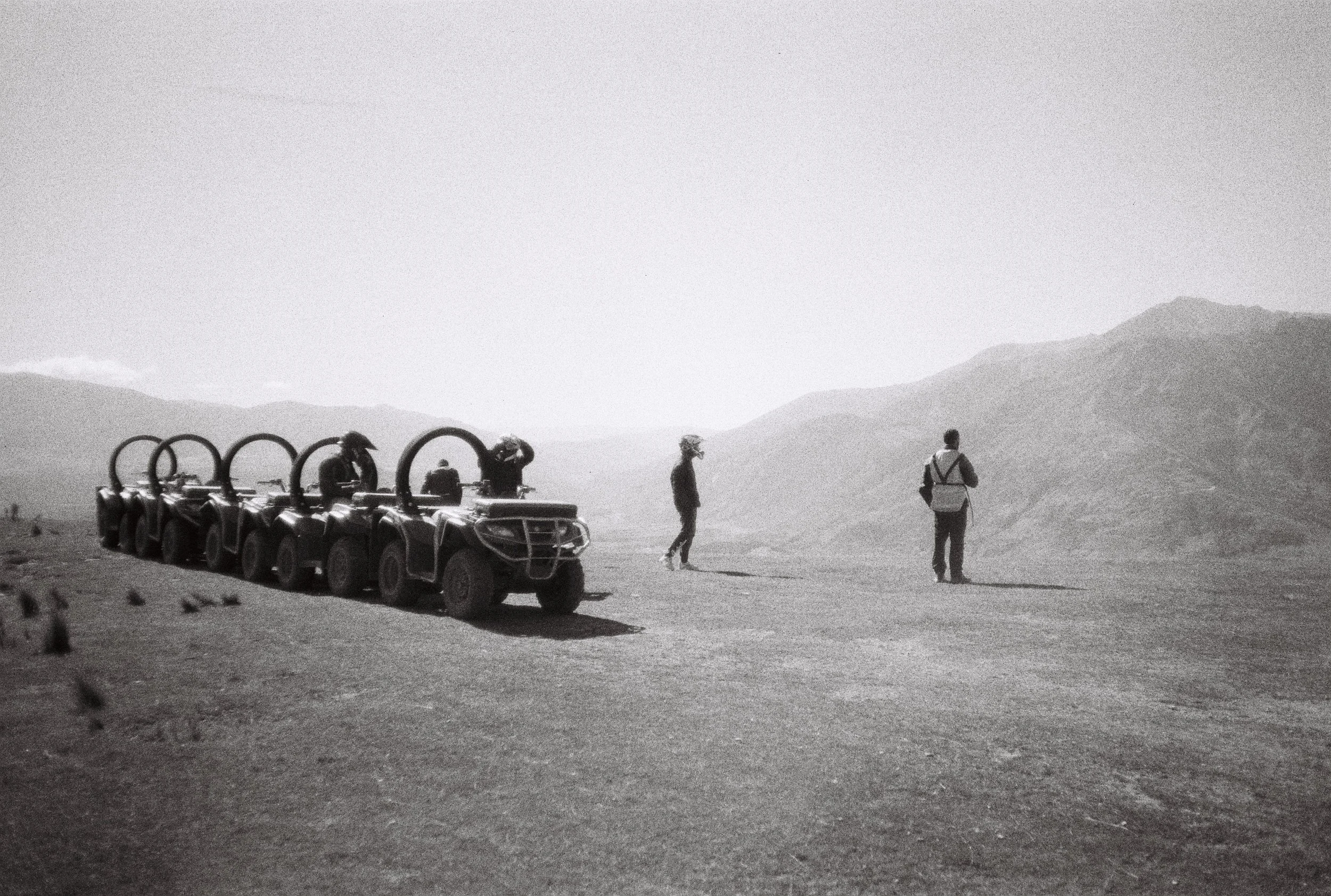 Groupe de personnes au milieu d'un désert, avec des véhicules tout-terrain alignés, montagnes à l'horizon, en noir et blanc.