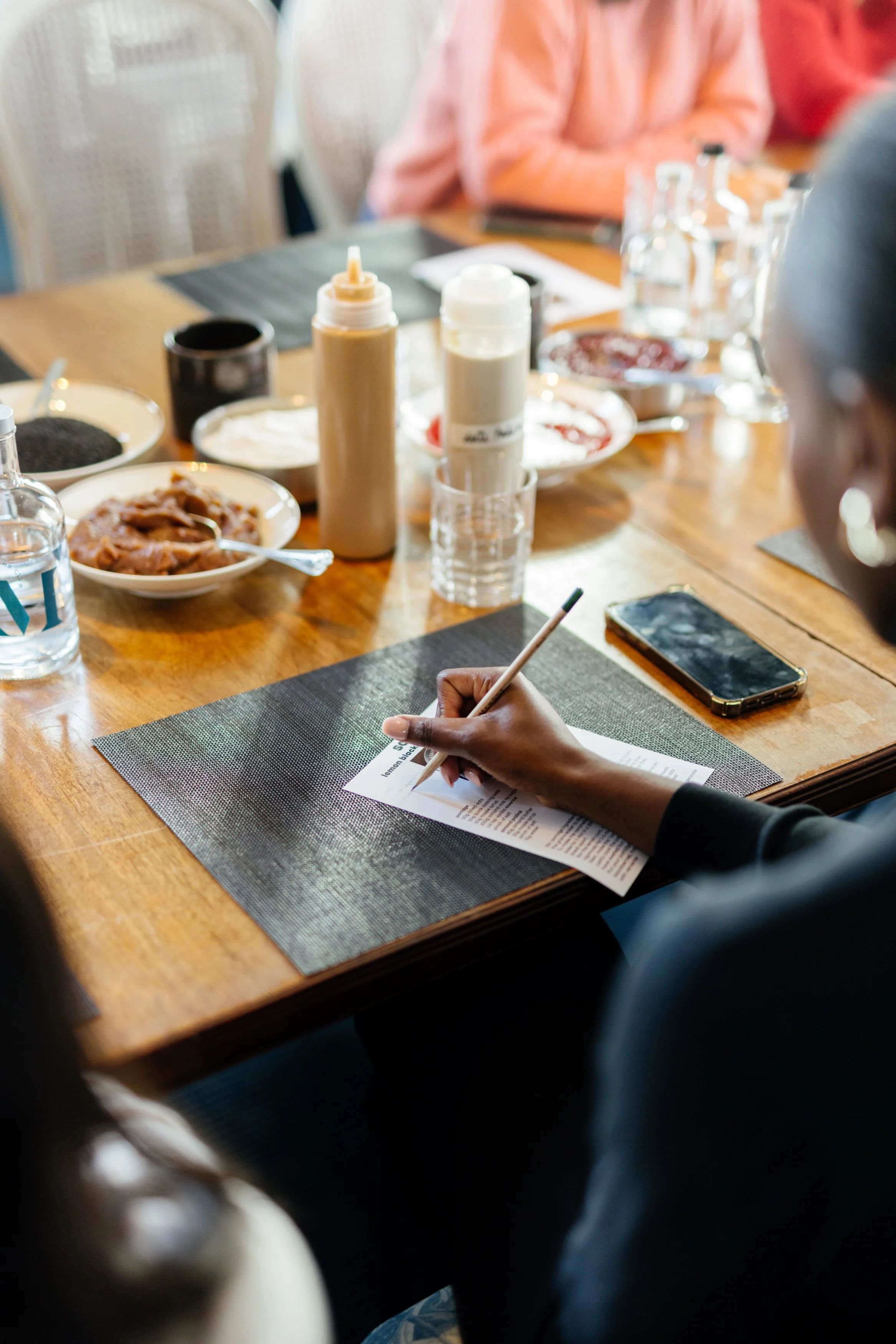 Une personne écrit sur une feuille à une table de restaurant avec plusieurs plats et boissons, un téléphone portable à côté.