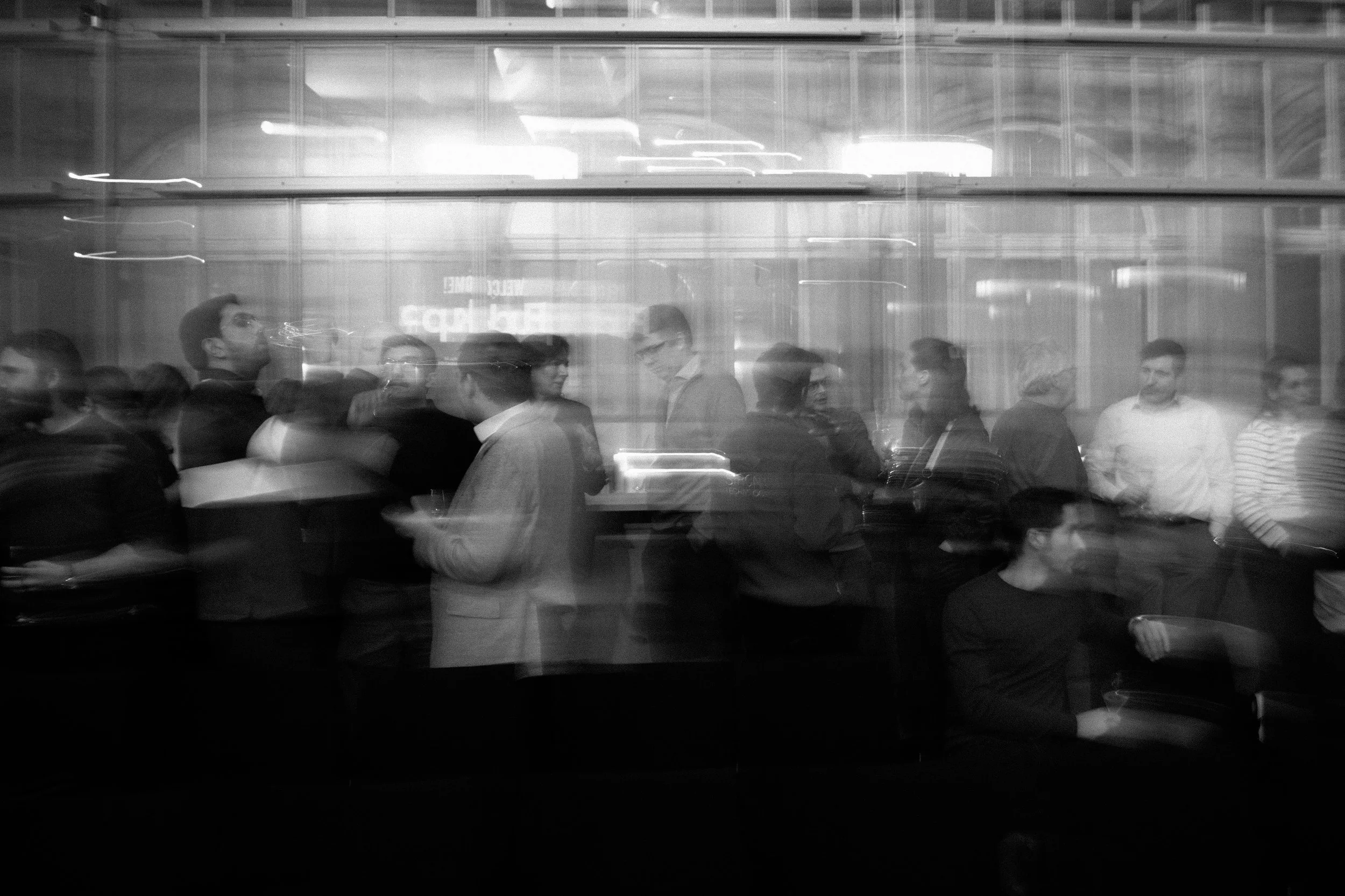 Groupe de personnes dans une file d'attente ou un rassemblement dans un espace intérieur, visibles à travers une vitre, avec un effet de flou lié au mouvement en noir et blanc.