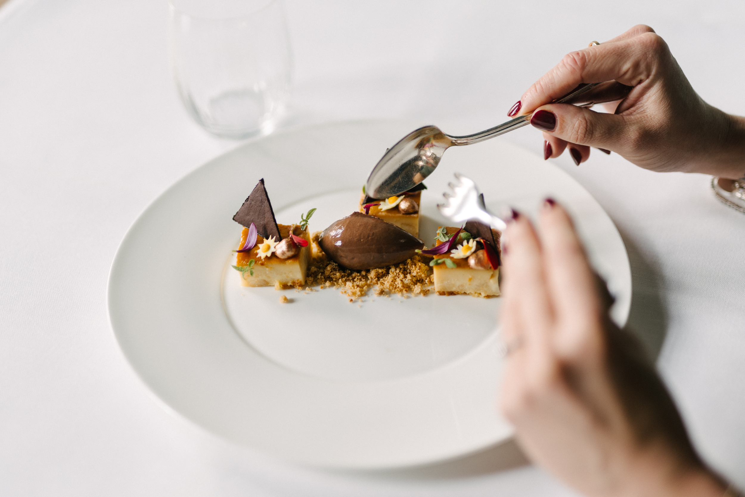 Une personne tient une cuillère au-dessus d'un dessert sophistiqué décoré de fleurs, de feuilles et de morceaux de chocolat sur une assiette blanche.