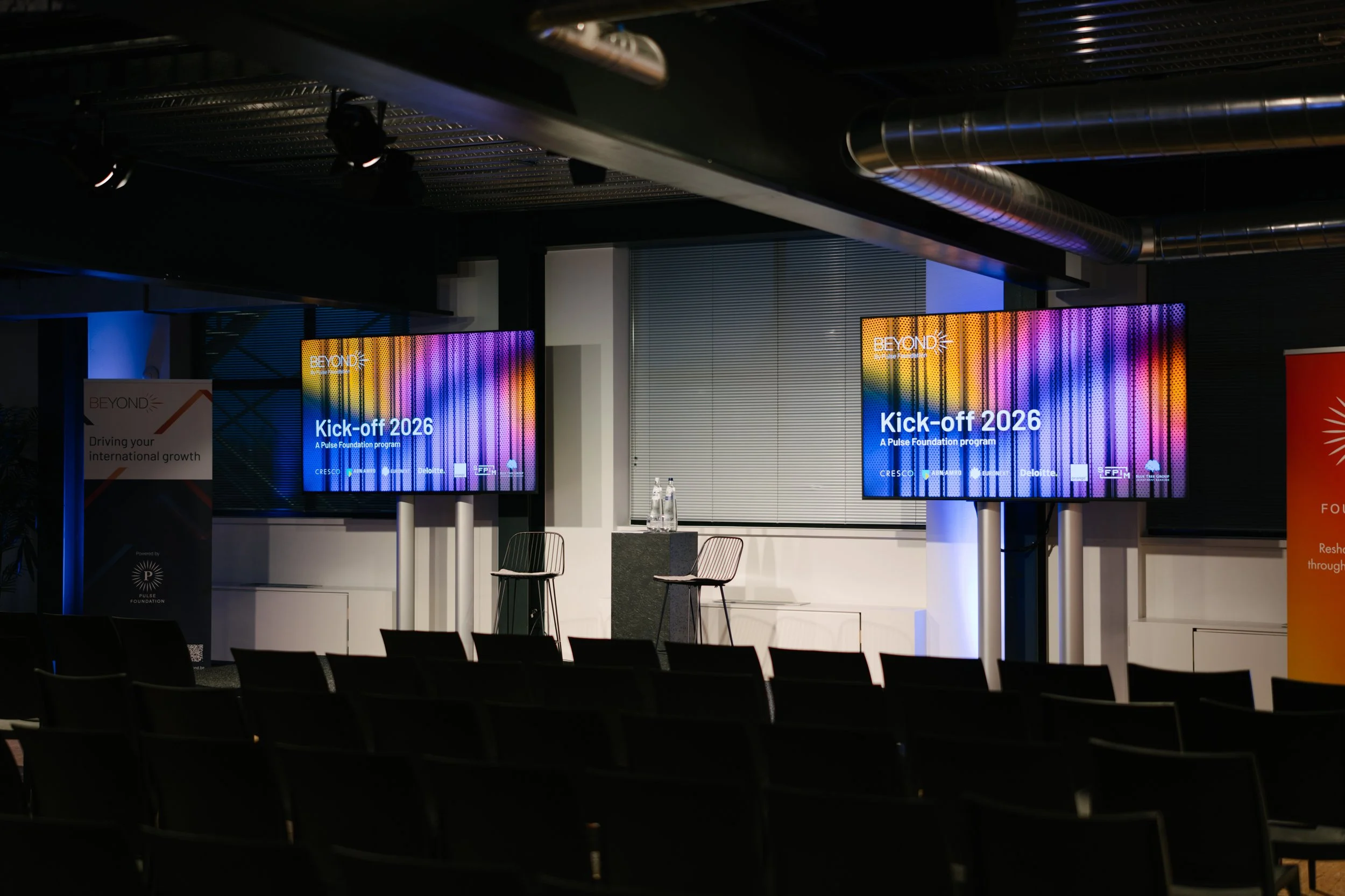 Salle de conférence avec deux écrans affichant 'Kick-off 2026', chaises vides, deux bouteilles d'eau sur une table, éclairage tamisé, décor moderne.