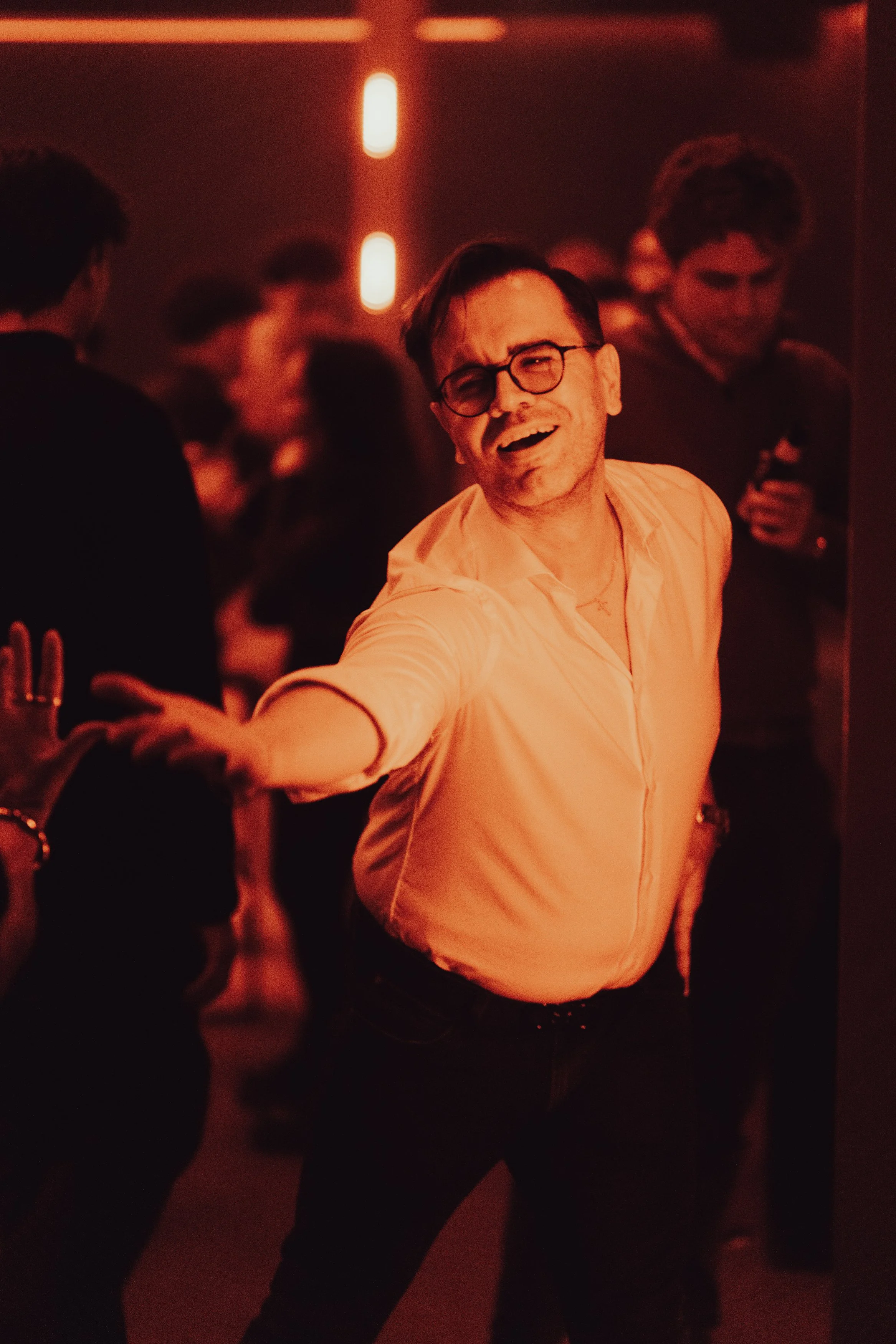 Homme avec lunettes, chemise claire, souriant et tendant la main dans une ambiance chaleureuse et sombre, avec des personnes en arrière-plan