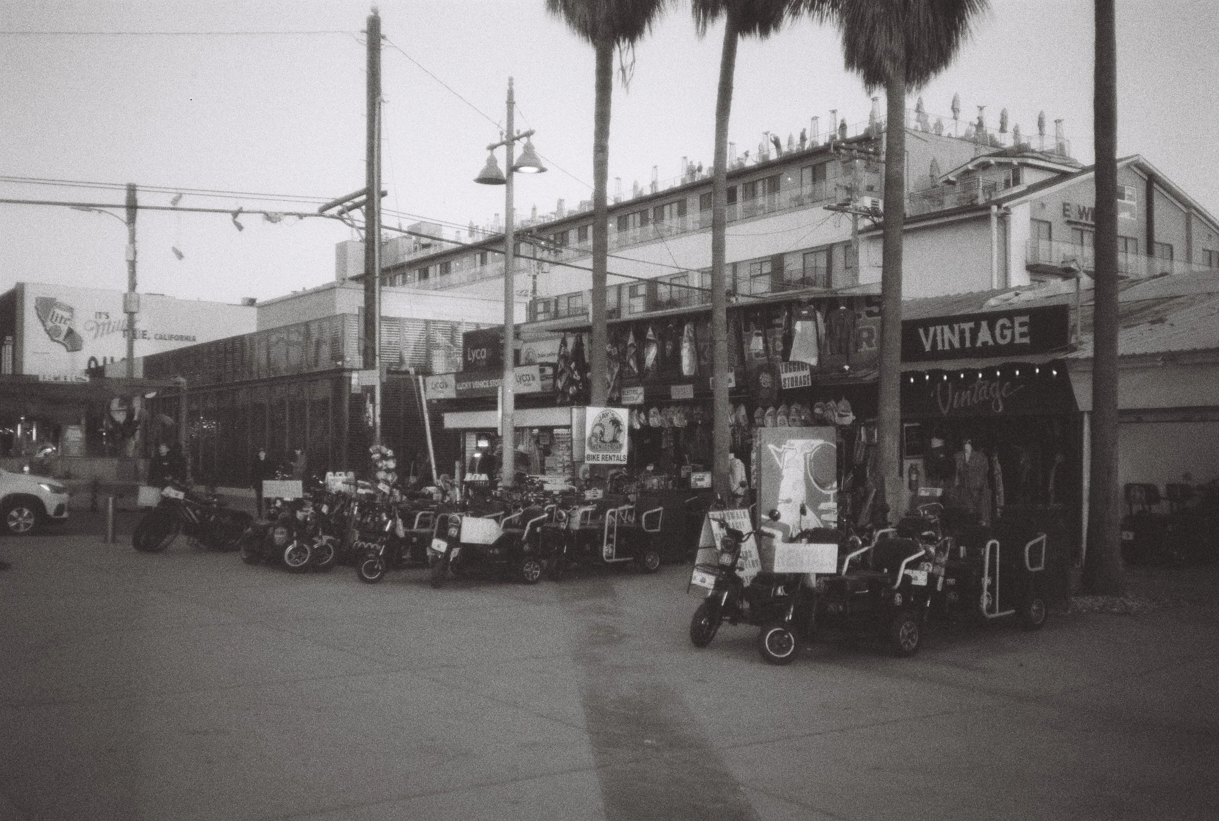 Une rue avec des motos et vélos électriques garés devant des boutiques, dont une boutique vintage, avec des palmiers et un immeuble résidentiel en arrière-plan.