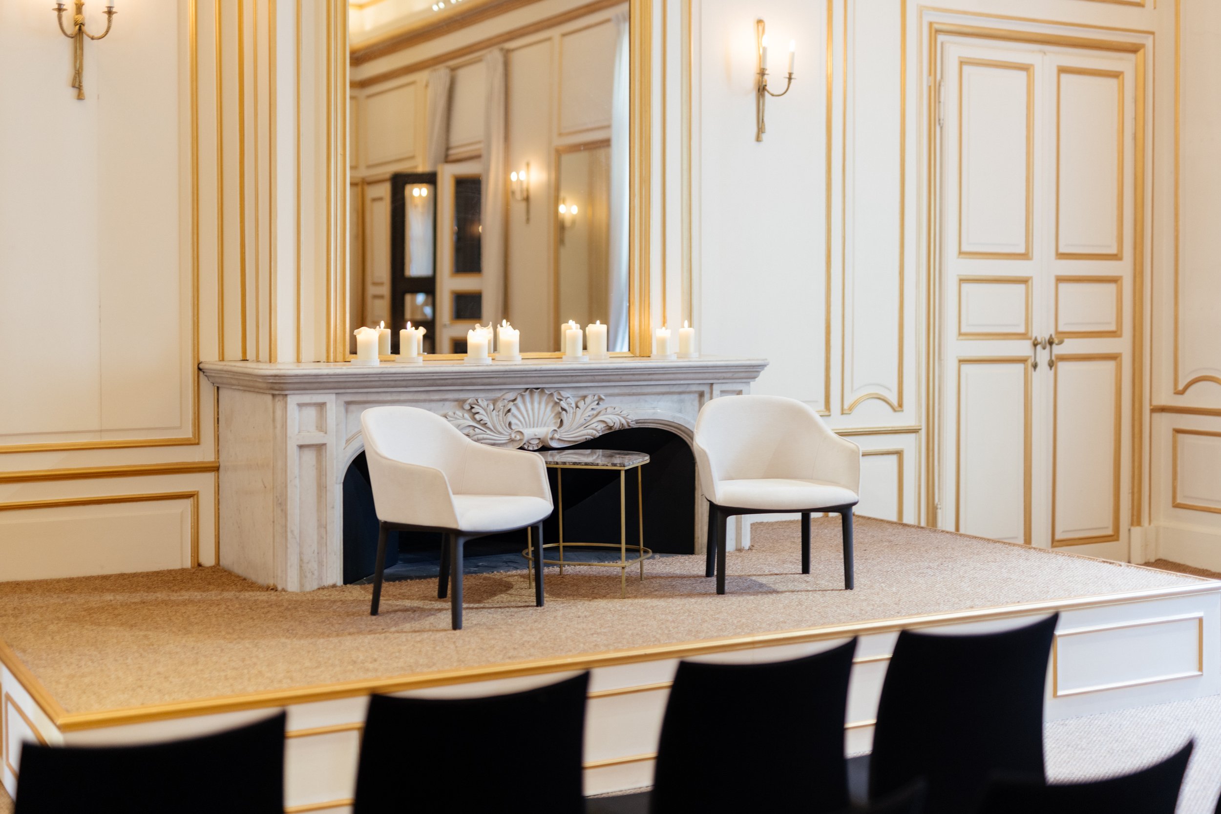 Une scène de salon avec deux fauteuils blancs, une cheminée en marbre décorée de bougies, un grand miroir, des murs ornementés en bois clair, et un tapis beige.