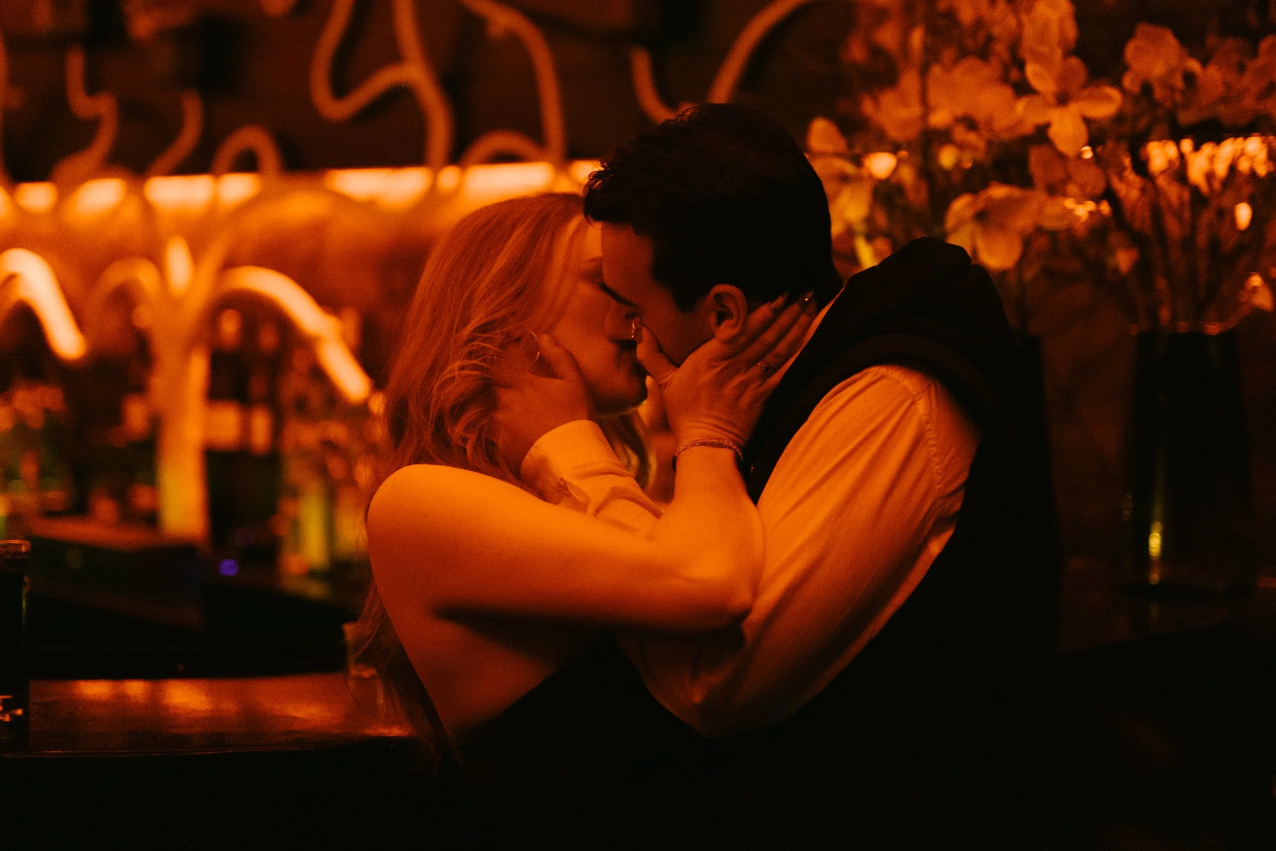 Un couple s'embrasse tendrement dans un bar ou un restaurant, avec un fond lumineux d'éclairage chaud et de fleurs décoratives.
