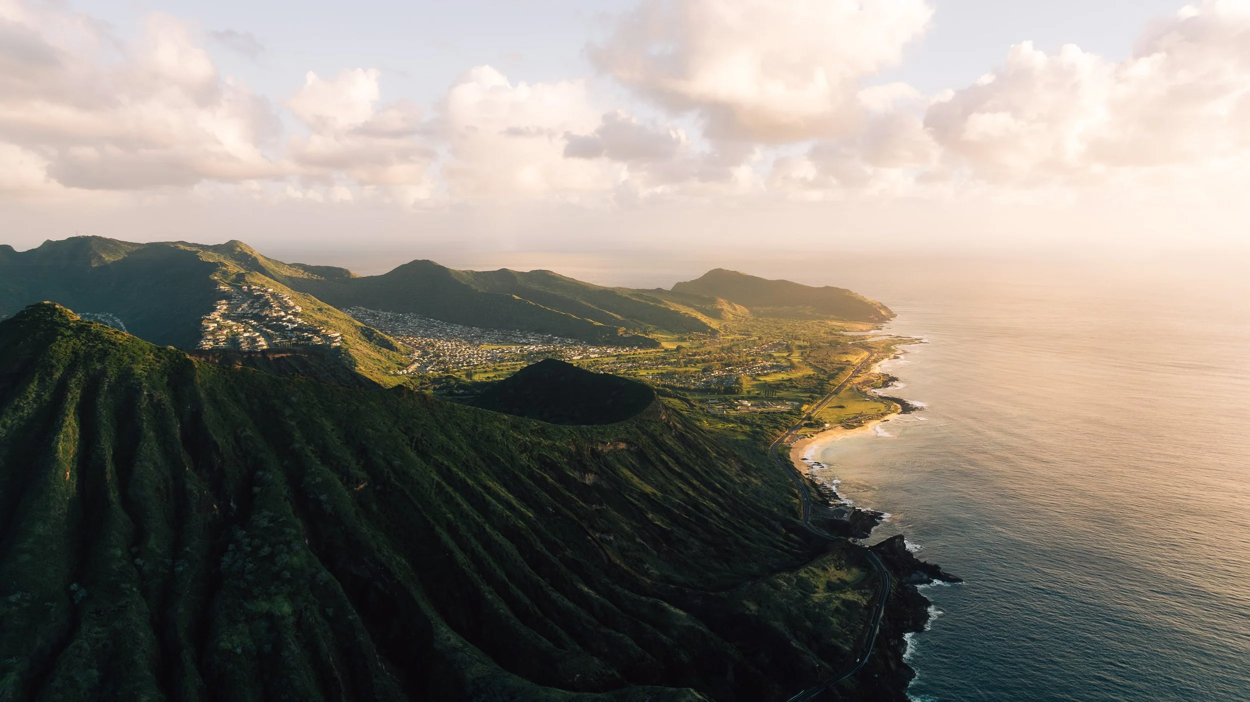 Paysage côtier avec montagnes verdoyantes, une ville, et l'océan au coucher du soleil.
