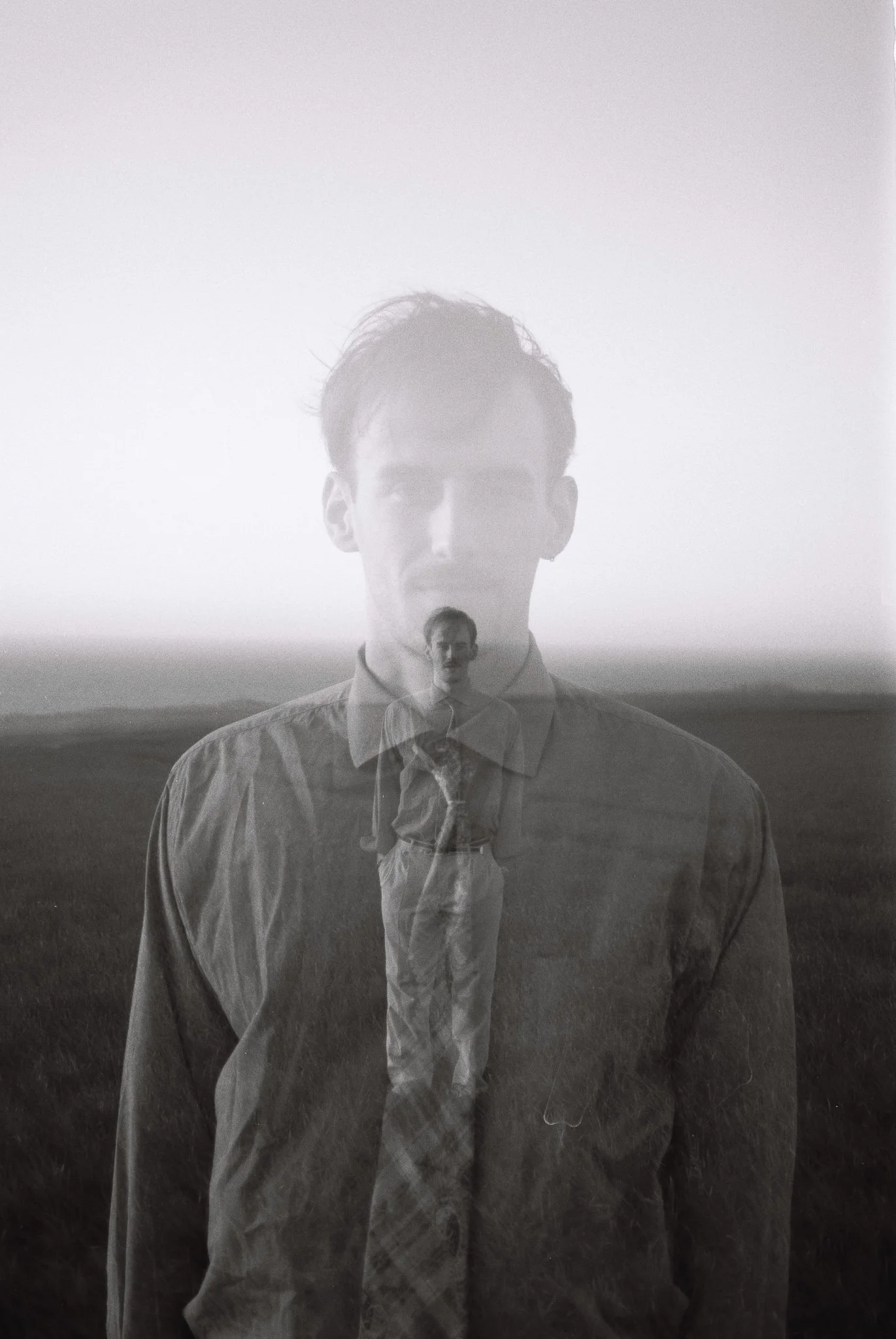Une double exposition en noir et blanc d'un homme en chemise avec cravate, superposé à l'image d'un paysage de campagne. La première image se concentre sur son visage, la seconde sur son corps avec le paysage en arrière-plan.