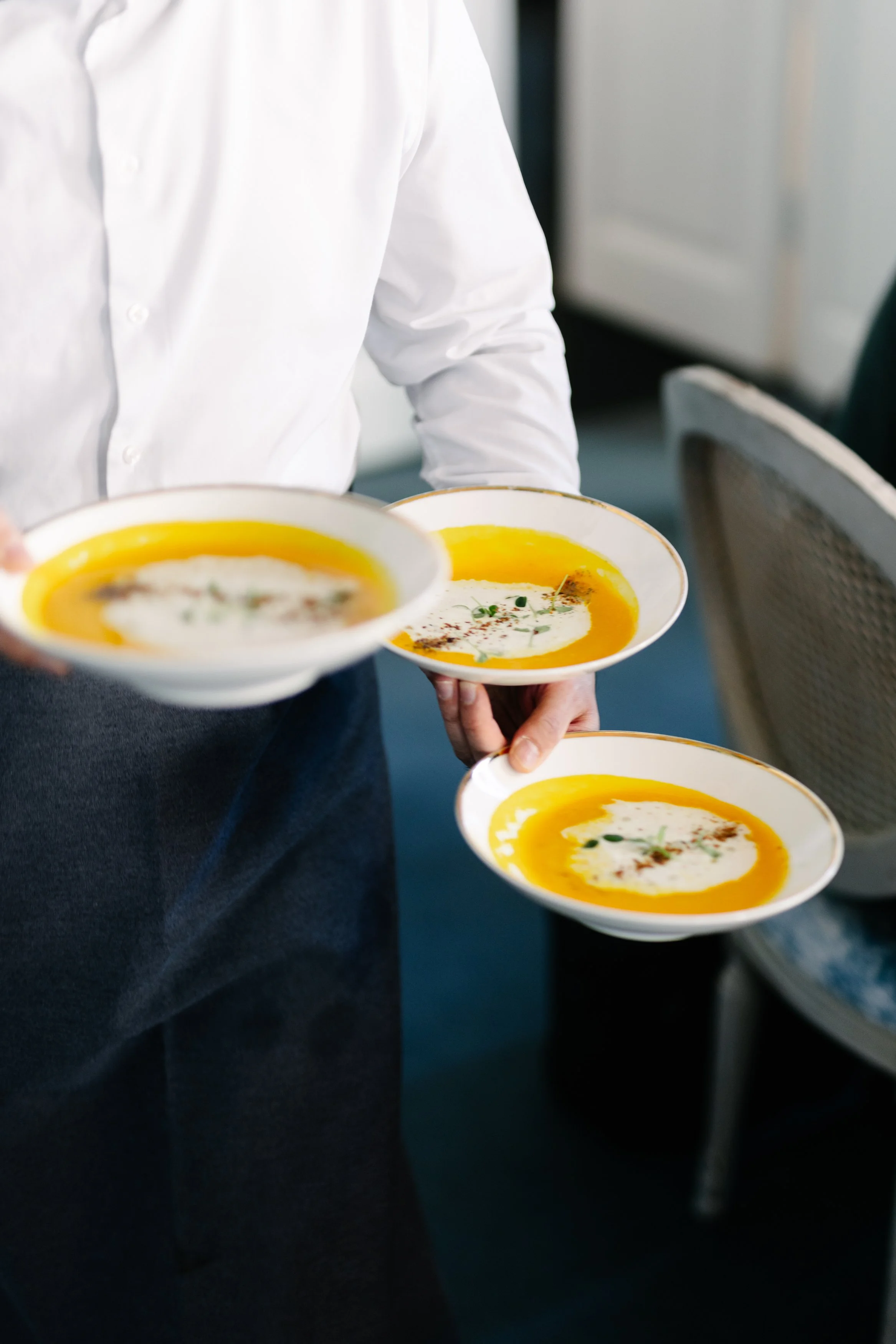 Une personne en chemise blanche sert des soupes de légumes dans de la vaisselle blanche avec une bande dorée.