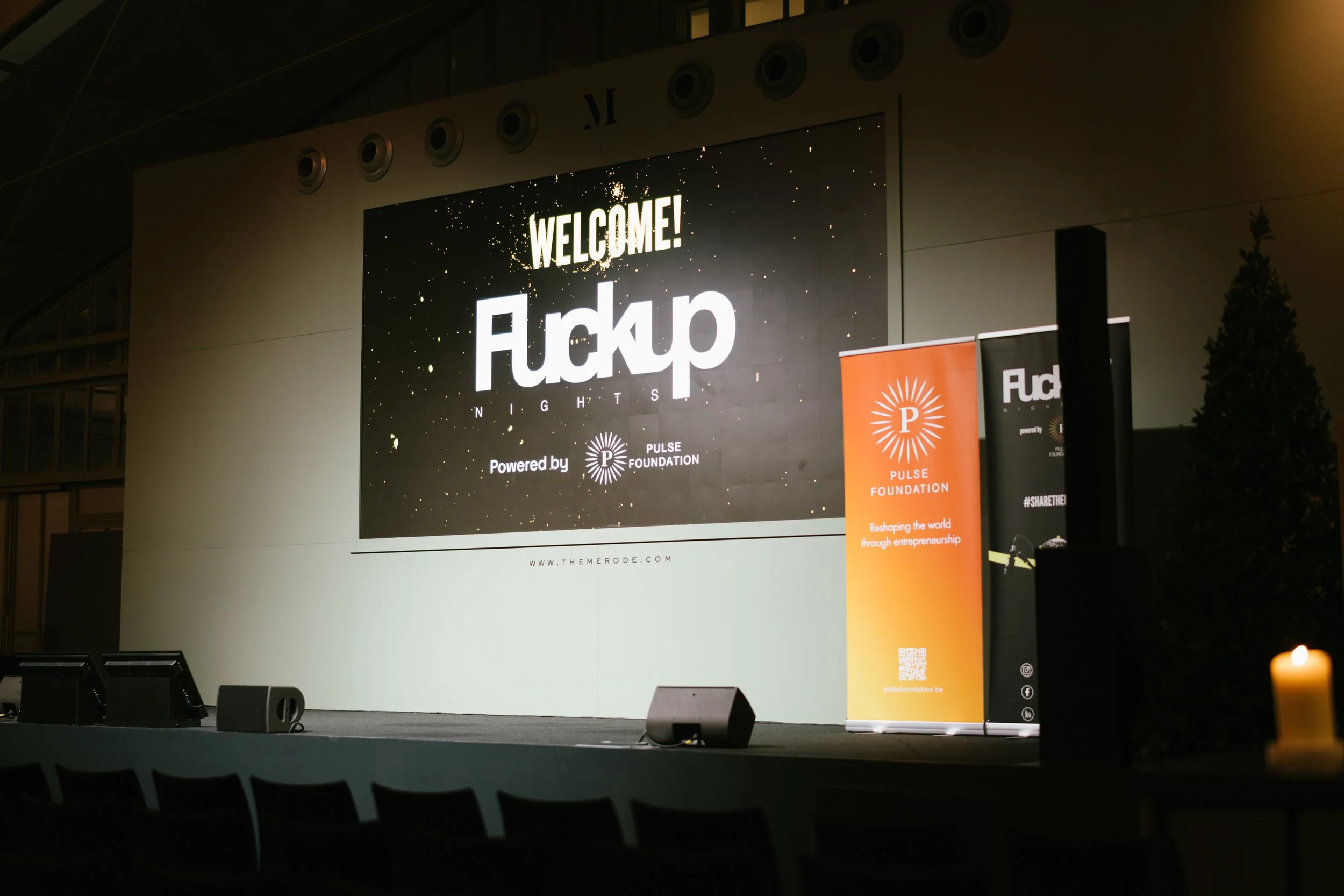 Une scène d'événement avec un grand écran affichant 'WELCOME! Fuckup Nights' et un bouton Power Foundation. Deux banderoles, l'une orange avec le logo de Pulse Foundation et l'autre noire, sont placées à côté de l'écran. Il y a aussi un chandelier av