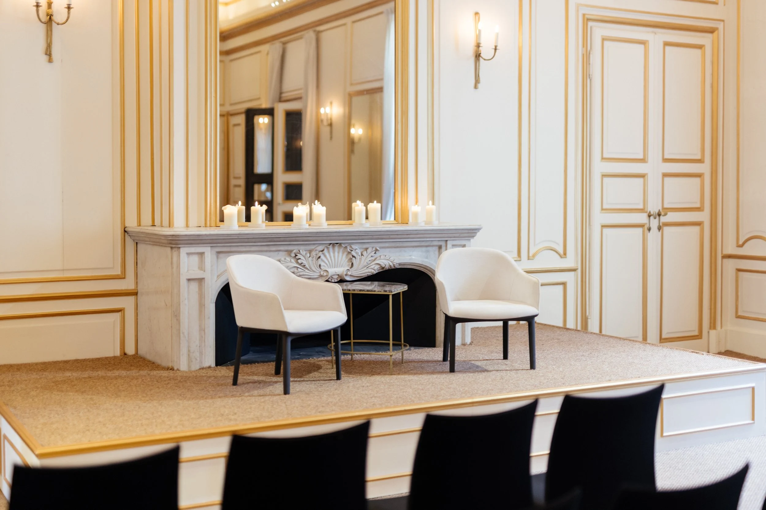 Une scène intérieure élégante avec deux fauteuils blancs placés devant une cheminée en marbre décorée de bougies. La pièce est ornée de boiseries dorées et de murs crème, créant une atmosphère sophistiquée.