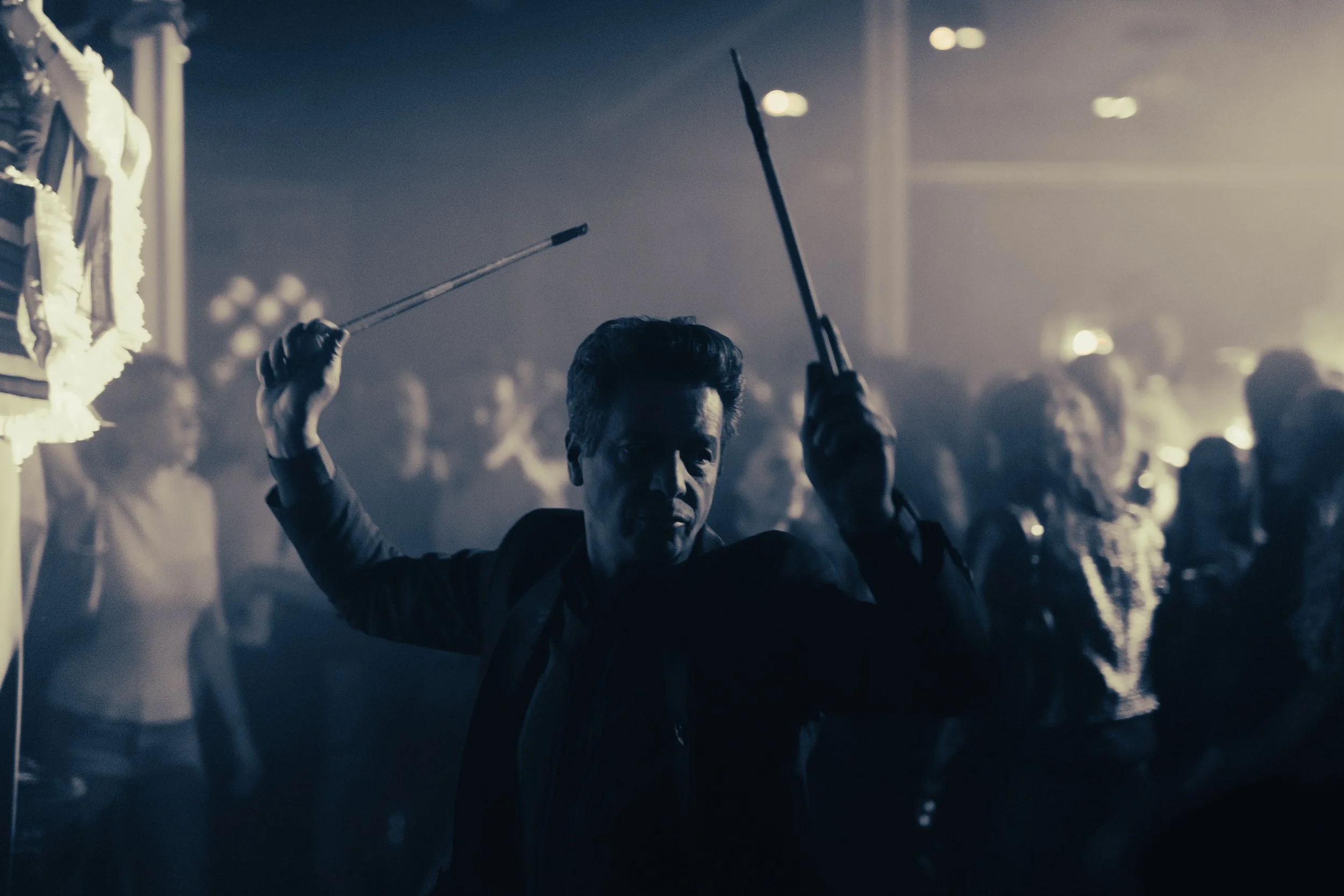 Un homme en costume noir dansant dans une foule sombre avec des lumières tamisées, tenant deux bâtons ou épées.