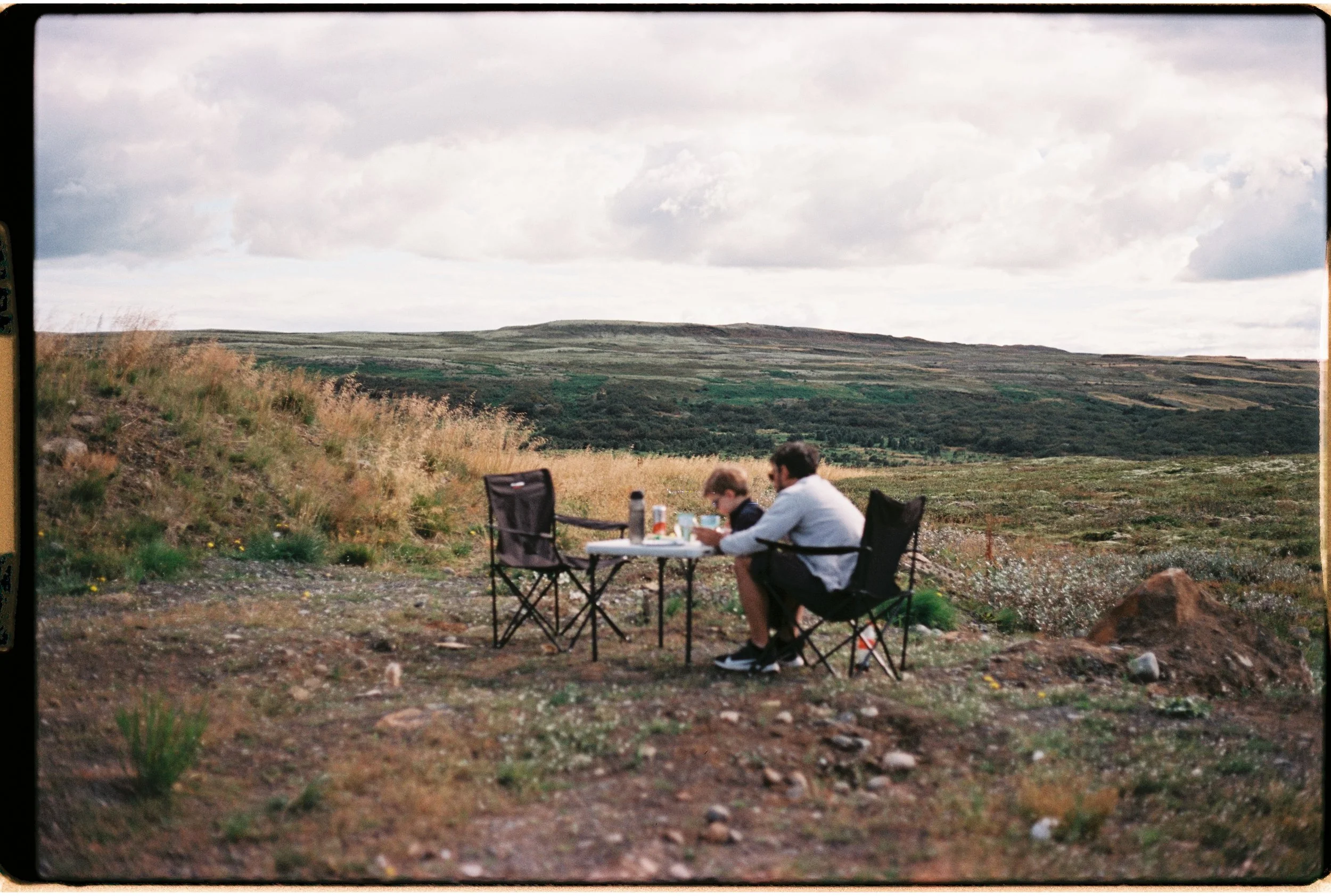 Deux personnes assises à une table pliante en plein air dans un paysage de campagne avec des collines verdoyantes, un ciel nuageux, et peu de végétation autour.