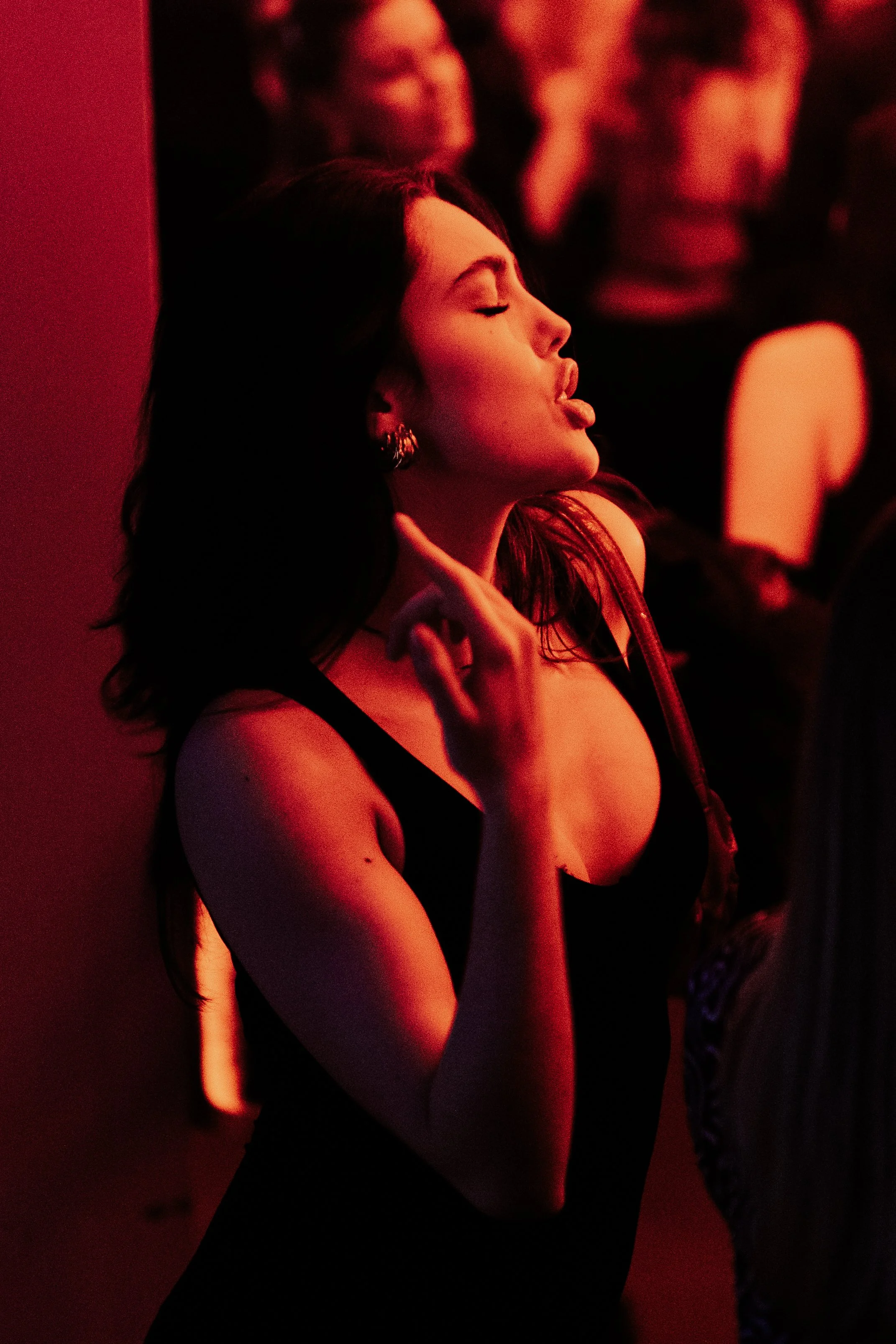 Une jeune femme avec des cheveux noirs, portant un haut noir, fait une grimace ou un mouvement de langue dans une ambiance sombre éclairée par une lumière rouge, dans une foule.