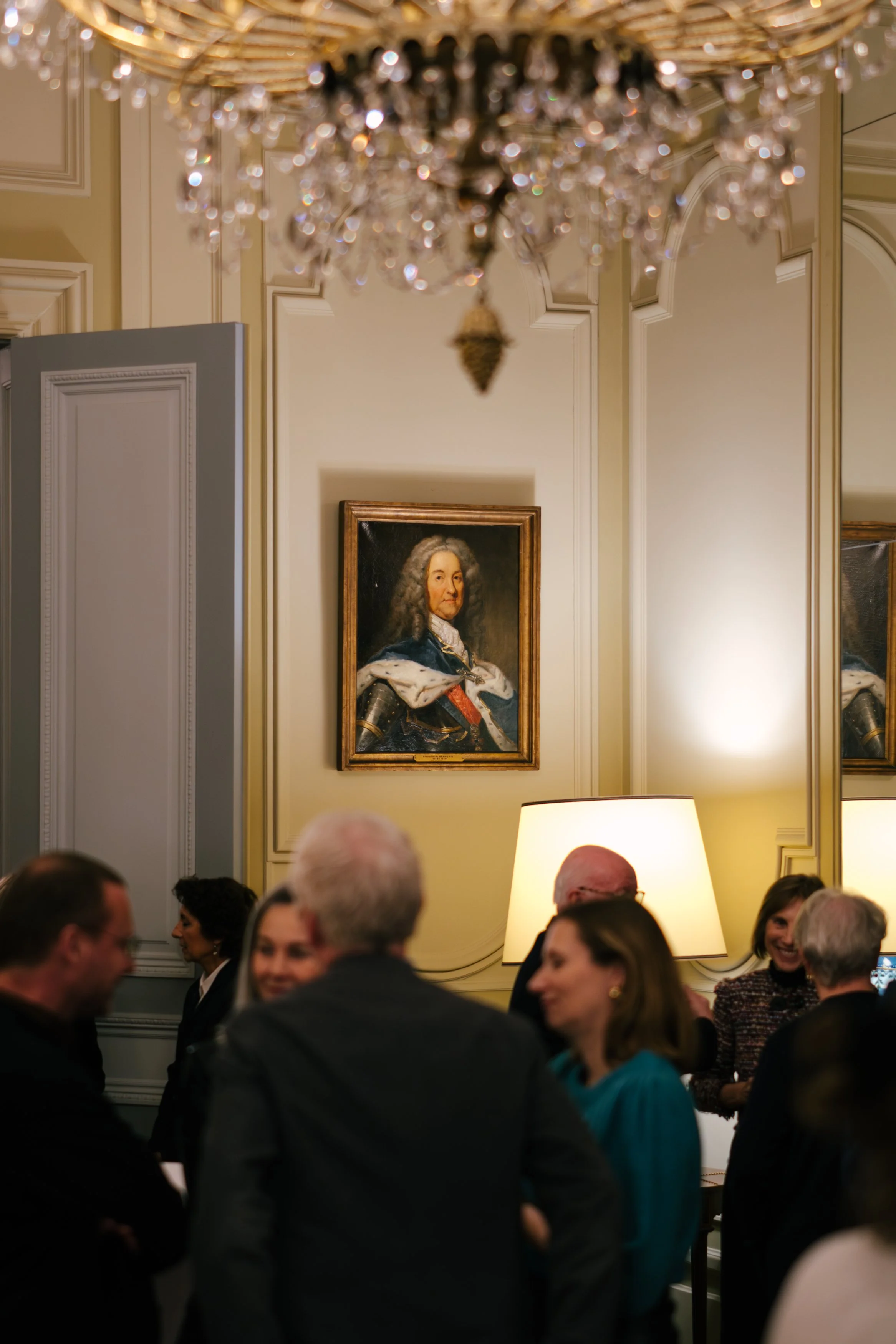 Une scène de rassemblement social dans une salle élégante, avec un plafond orné d'un chandelier scintillant, un portrait de style classique d'un homme en uniforme suspendu au mur, et des personnes discutant dans une ambiance chaleureuse et sophistiqu