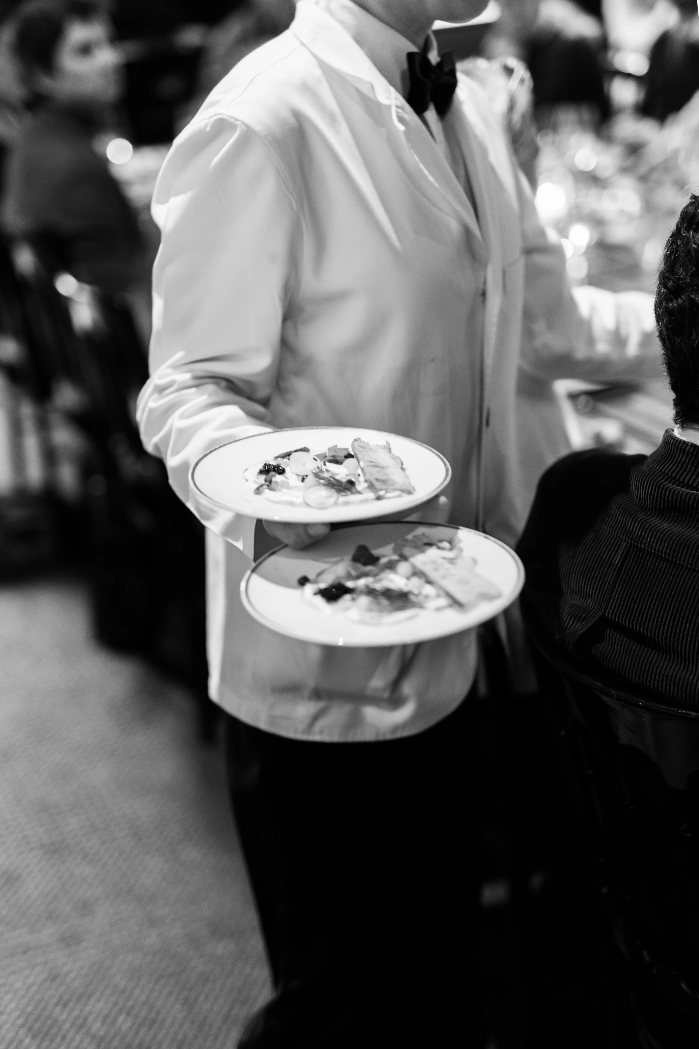 Un serveur en uniforme blanc avec un nœud papillon noir tenant deux assiettes de nourriture dans un restaurant