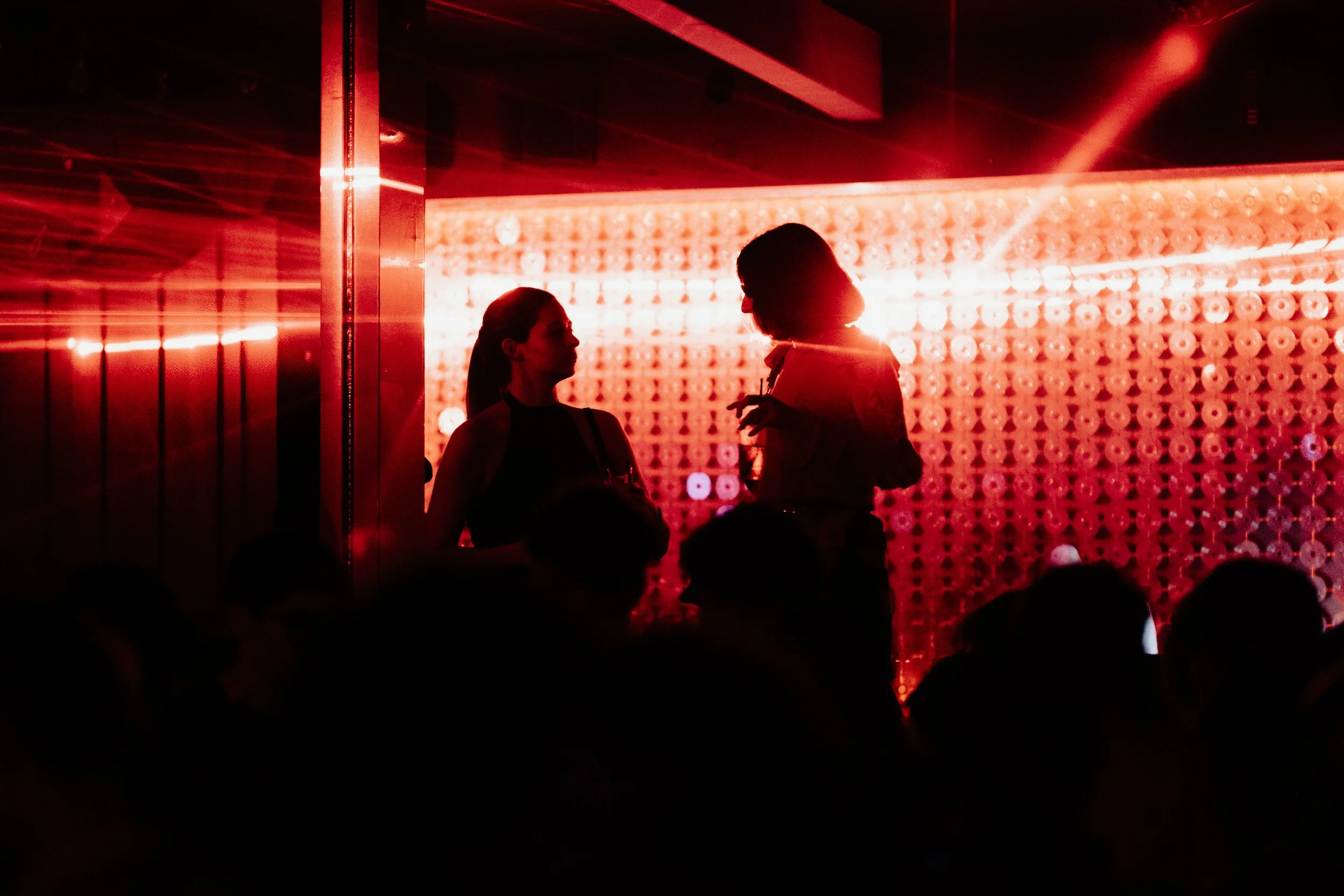 Deux femmes en silhouette discutent dans une ambiance rouge et sombre, avec un mur de lumières en arrière-plan, dans un lieu probablement une fête ou un club.