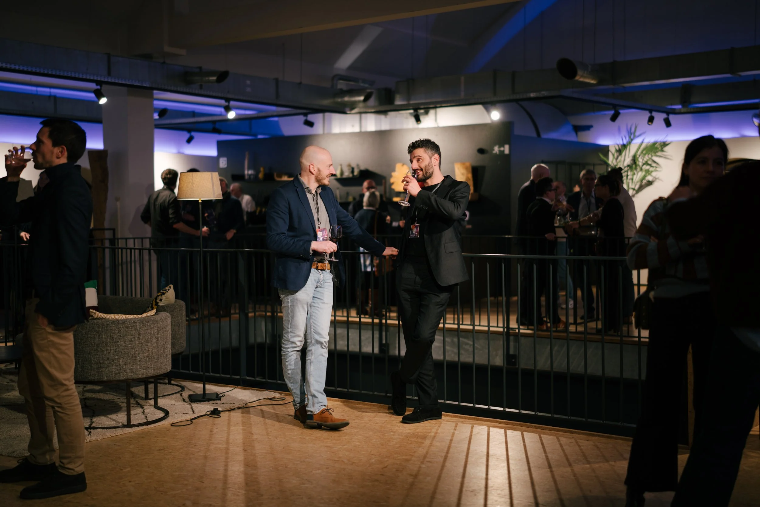 Deux hommes discutent et tiennent des verres à un événement social dans une salle moderne, entourés d'autres personnes qui socialisent, avec un éclairage d'ambiance.