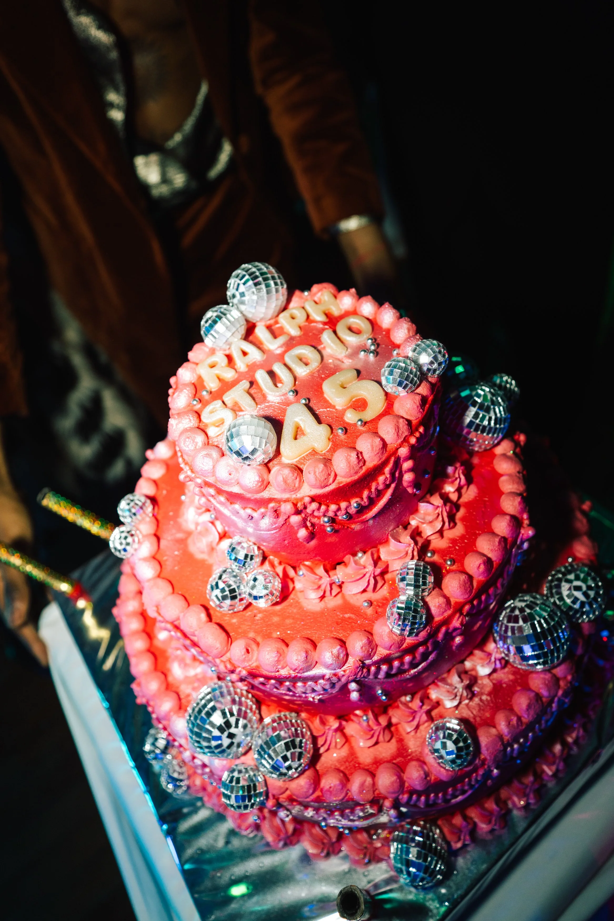 Gâteau d'anniversaire rose à trois étages décoré de disco ball en miroir et de petites fleurs, avec inscription en lettres blanches "RAPHAËL STUDIO C 4, S" en haut.