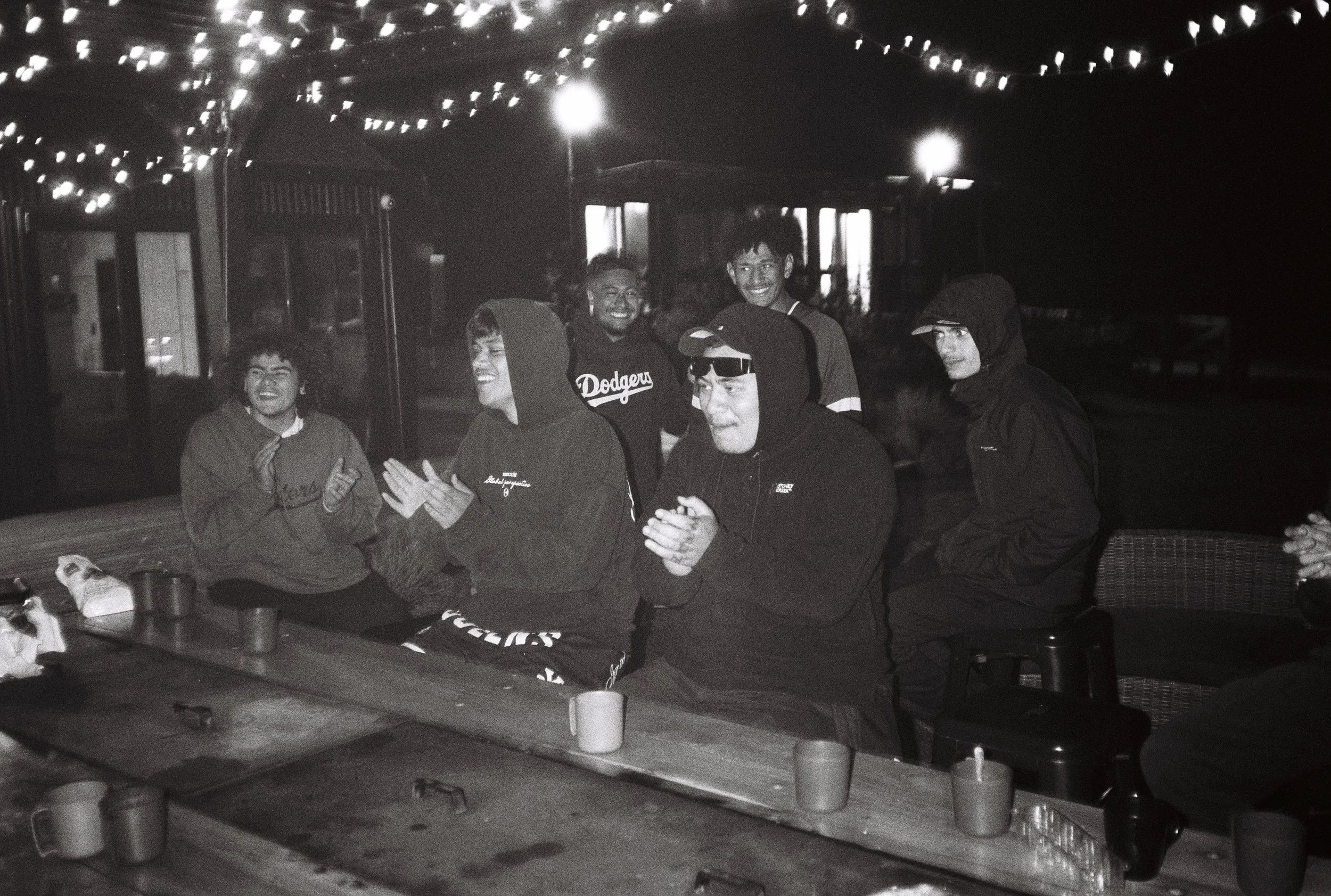 Groupe de personnes assises et debout sous un ciel nocturne, éclairées par des guirlandes lumineuses, en train de rire et d'applaudir lors d'un rassemblement en extérieur.