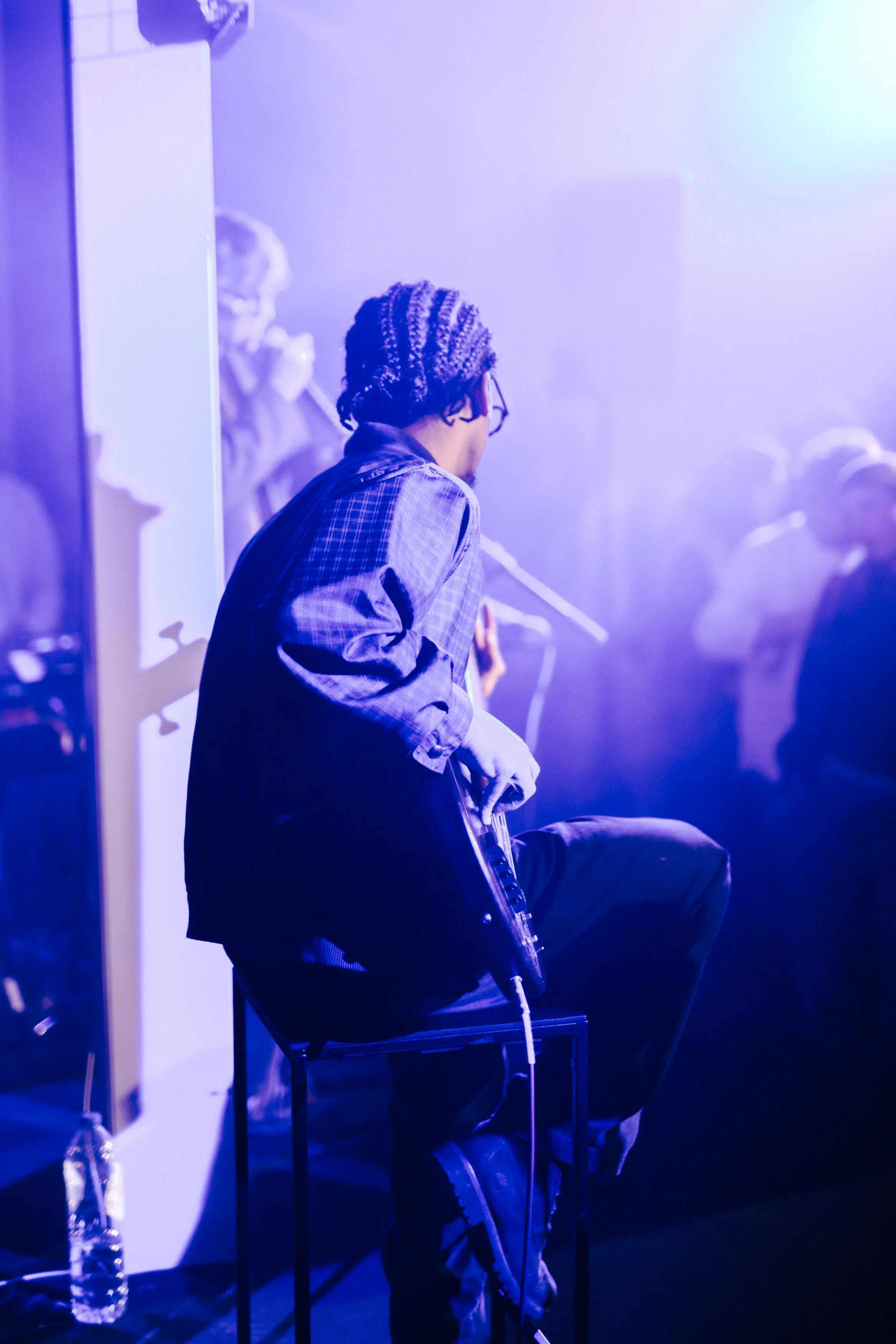 Un musicien joue de la guitare dans une ambiance de concert avec des lumières violettes et bleues, assis sur une chaise, portant des vêtements décontractés et des cheveux bouclés avec un bonnet.