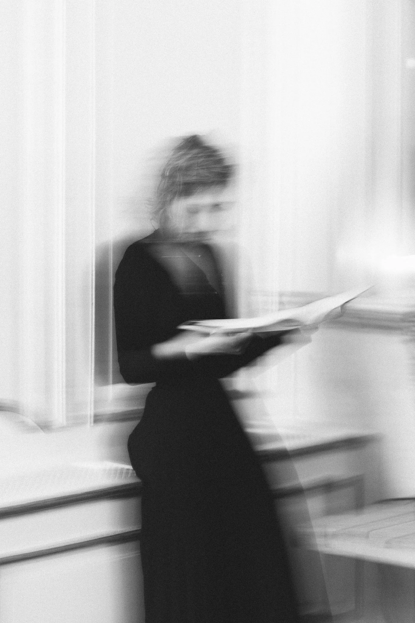 Une femme se tenant dans une pièce, tenant un livre ou un dossier, avec un effet de flou et de mouvement dans la photo.