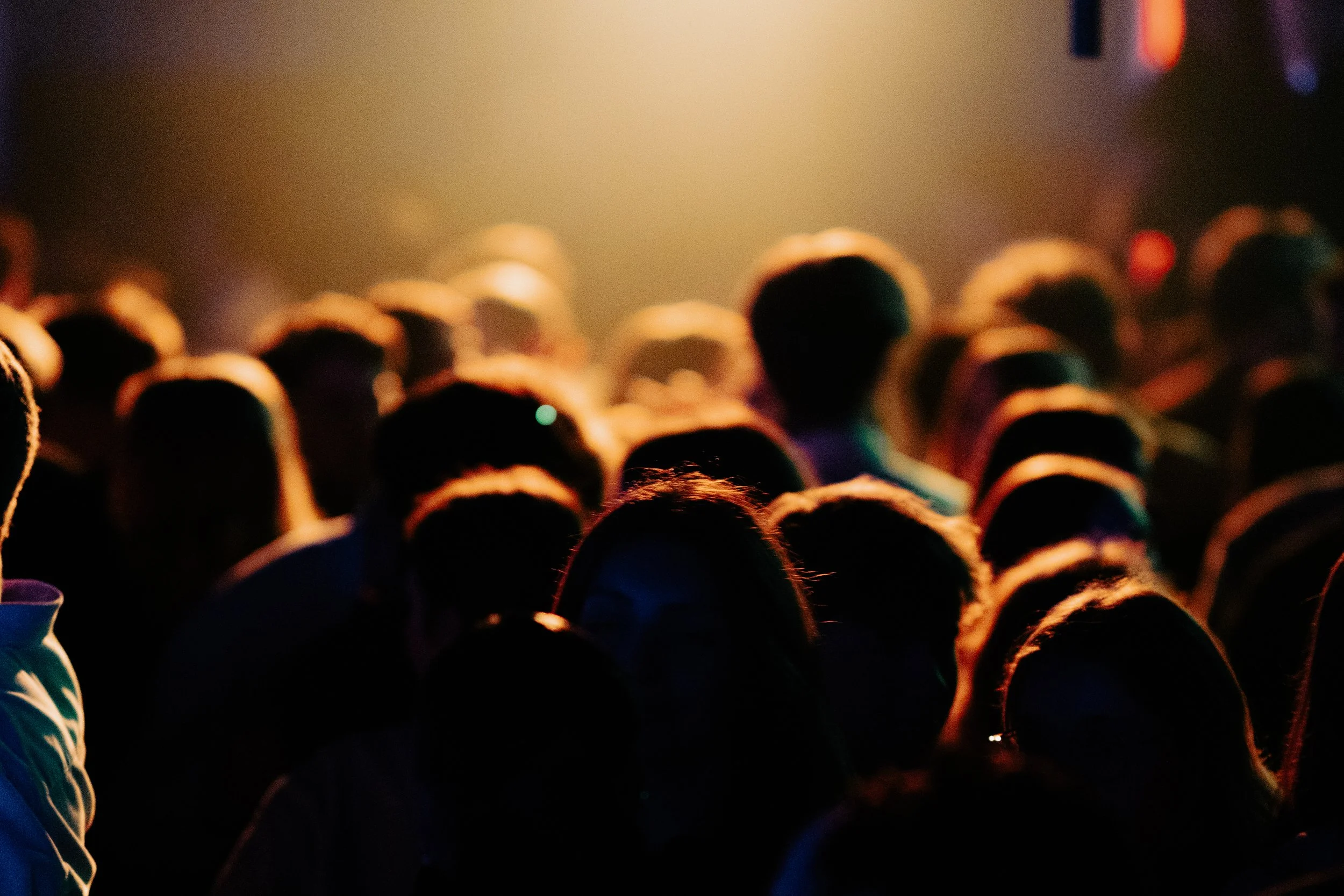Une foule de personnes au coucher du soleil, avec un éclairage chaud et des silhouettes noires, dans un espace intérieur ou extérieur.