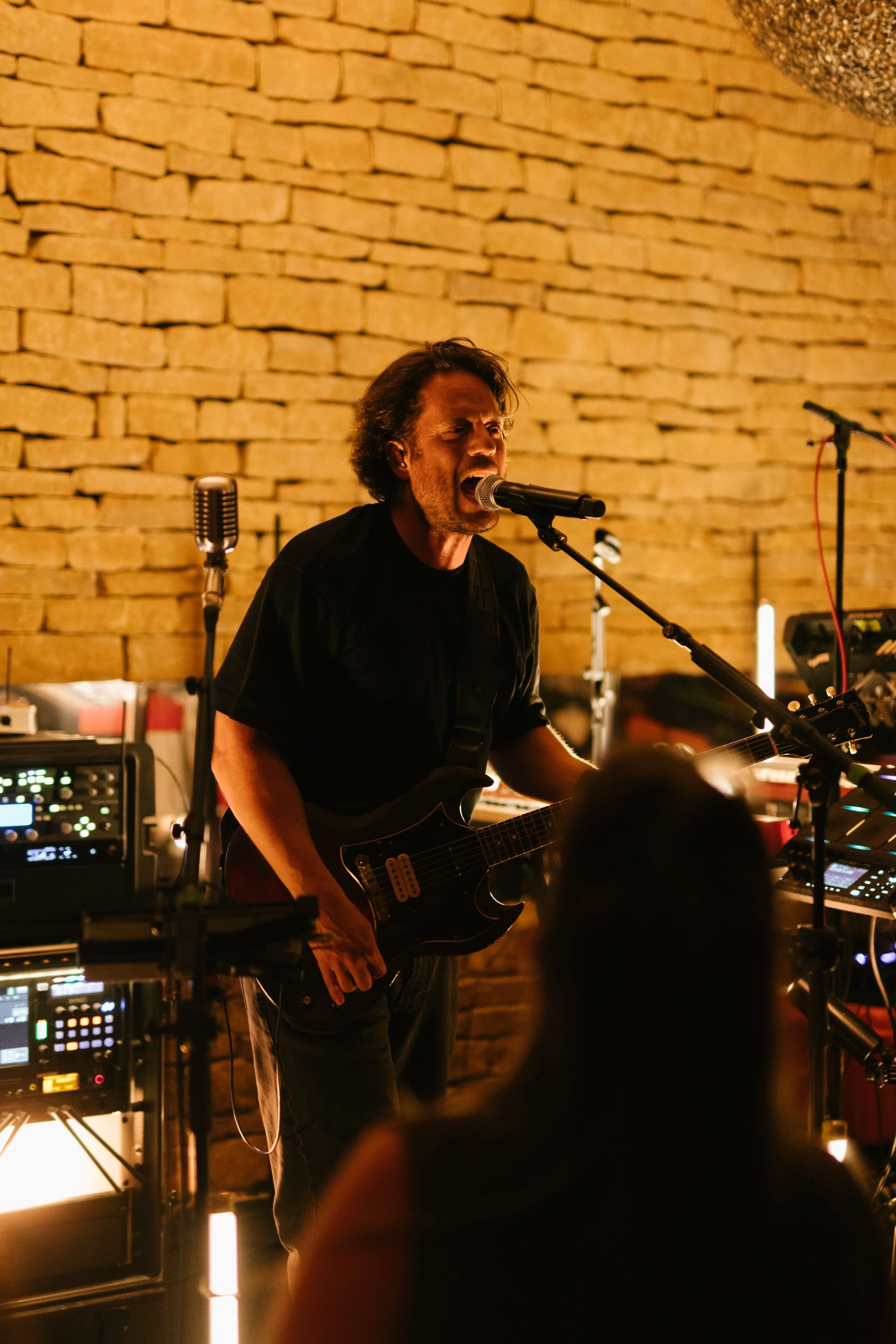 Un homme chante et joue de la guitare électrique lors d'une performance en intérieur, avec un mur en briques en arrière-plan, éclairé par une lumière chaleureuse.