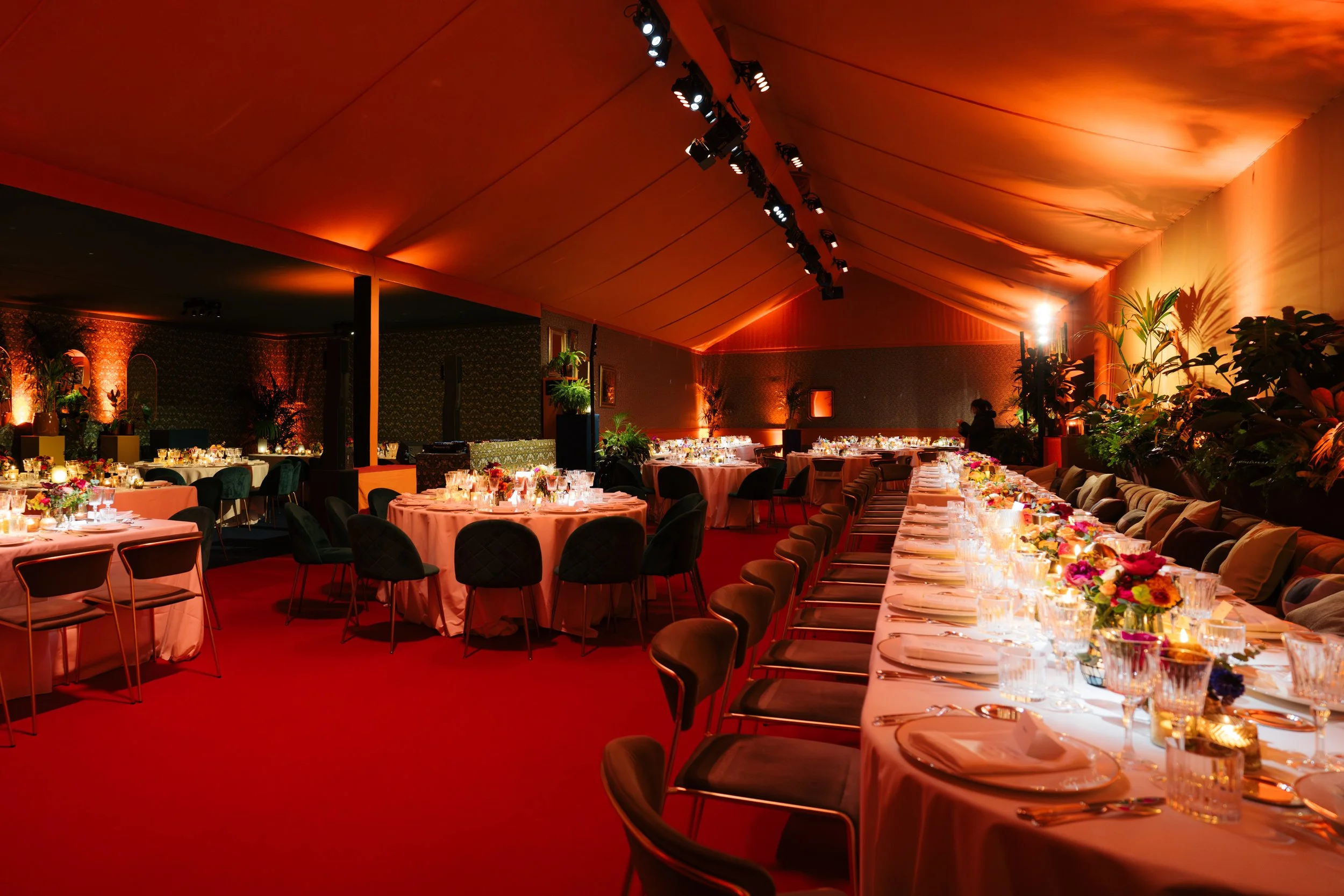 Une salle de banquet élégante décorée pour un événement, avec des tables rondes et une longue table rectangulaire, toutes ornées de nappes blanches, de bouquets de fleurs colorés et de bougies, avec un éclairage chaud et tamisé