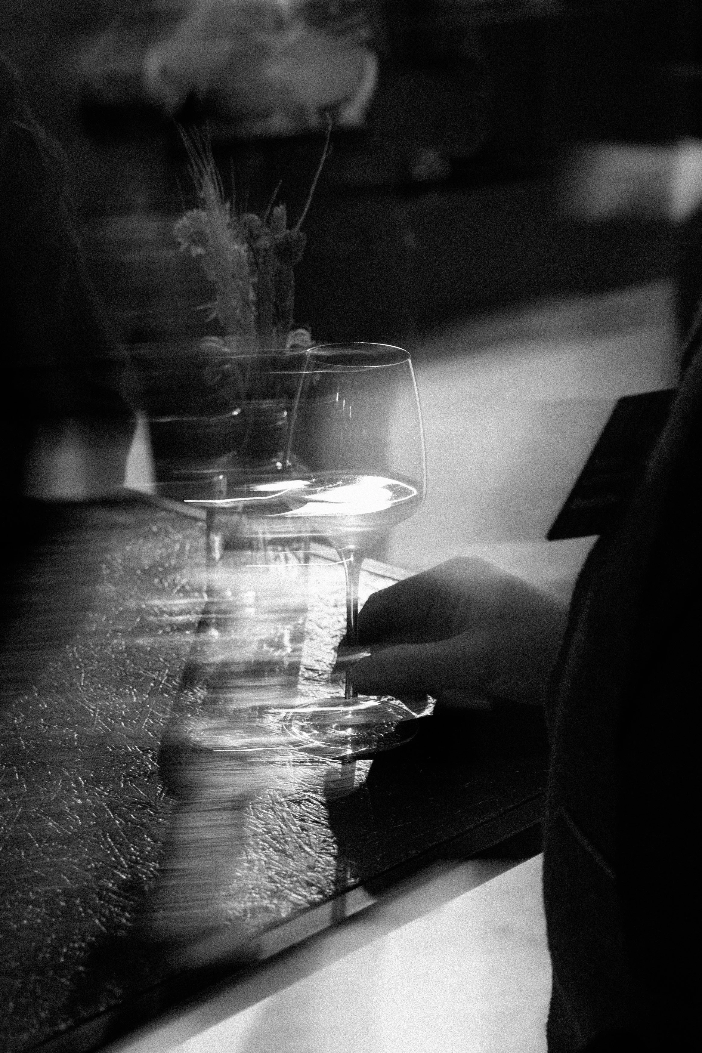 Une personne tenant un verre de vin blanc sur une table en bois, avec une vase contenant des fleurs en arrière-plan, en noir et blanc.