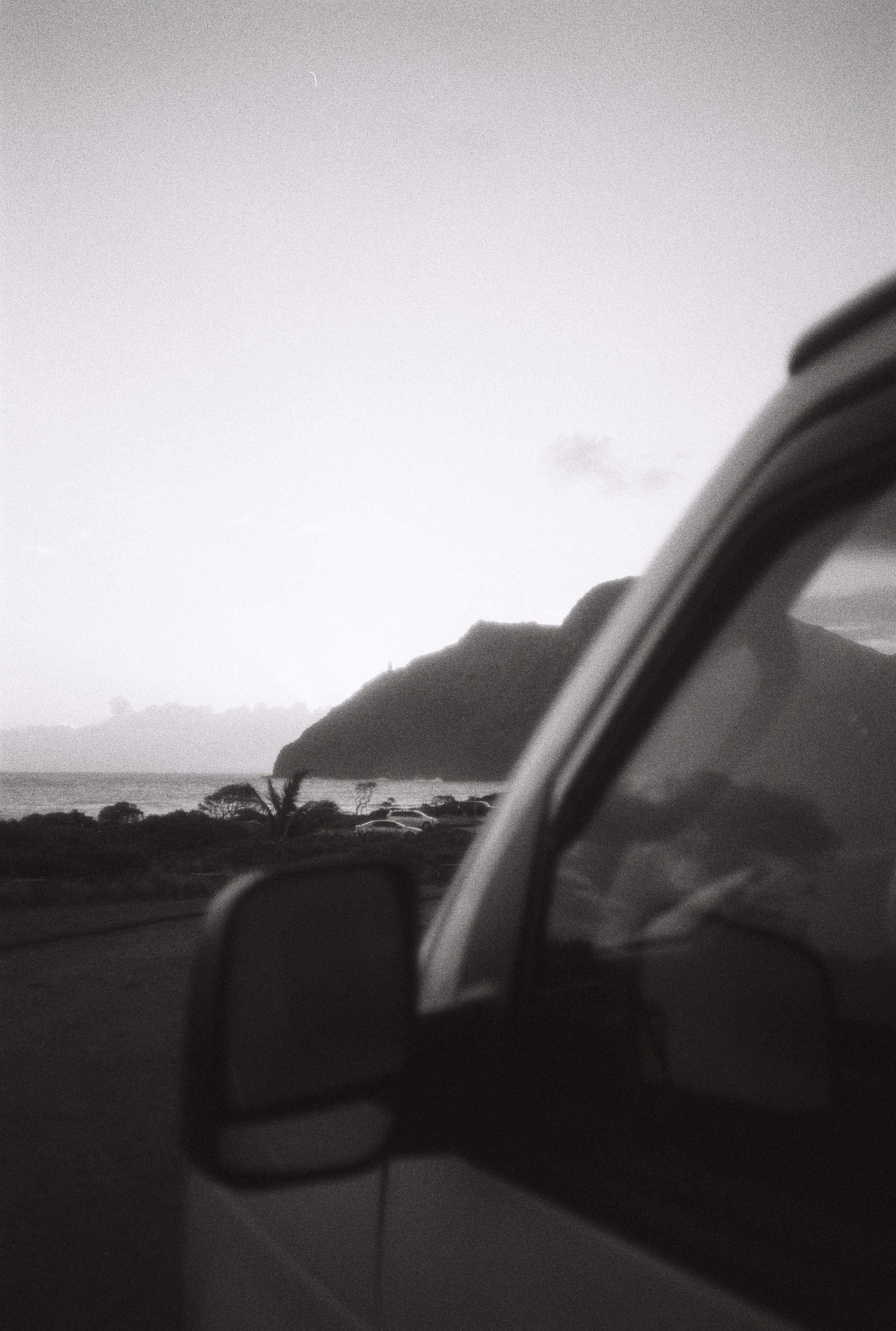 Une vue en noir et blanc d'une voiture avec un paysage côtier en arrière-plan, comprenant une mer, des collines, et quelques arbres, lors du crépuscule ou au lever du soleil.