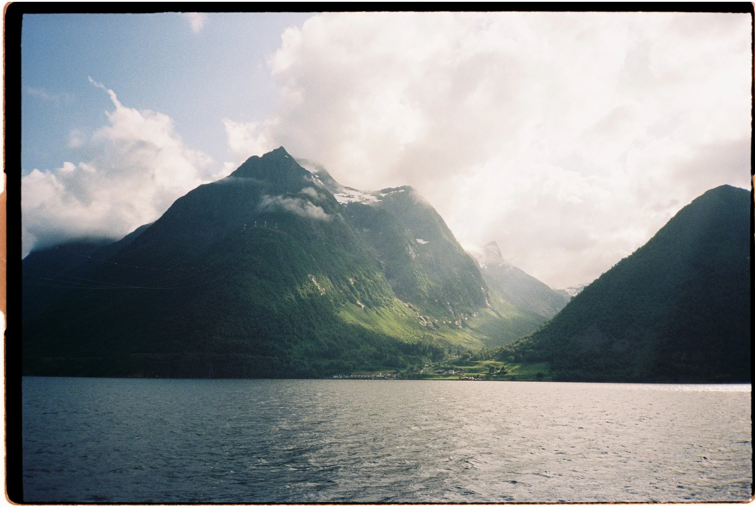 Paysage de montagnes verdoyantes au bord d'un lac sous un ciel partiellement nuageux.
