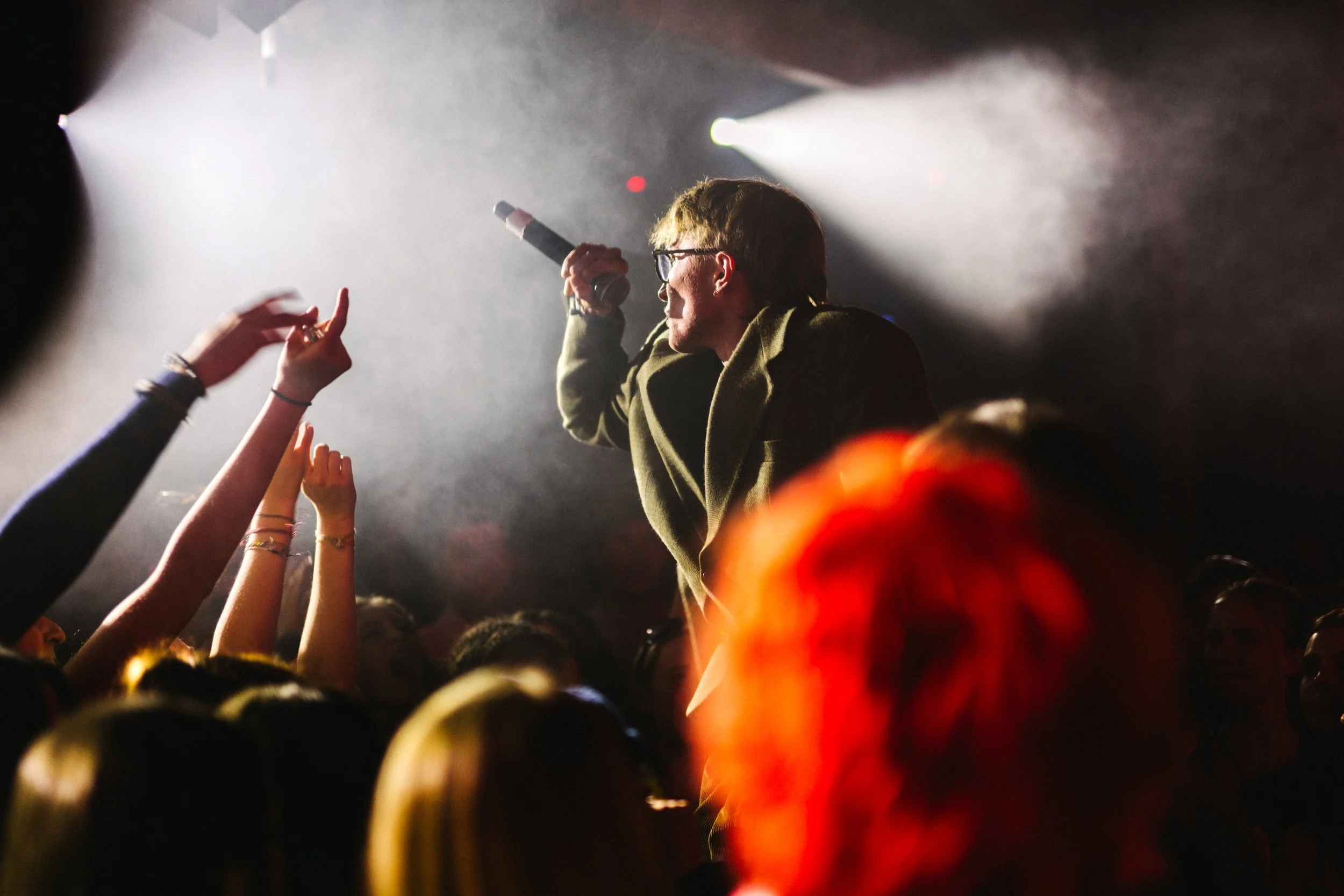 Un chanteur ou rappeur avec des lunettes, tenant un micro, se produit sur scène devant un public dans une lumière tamisée.