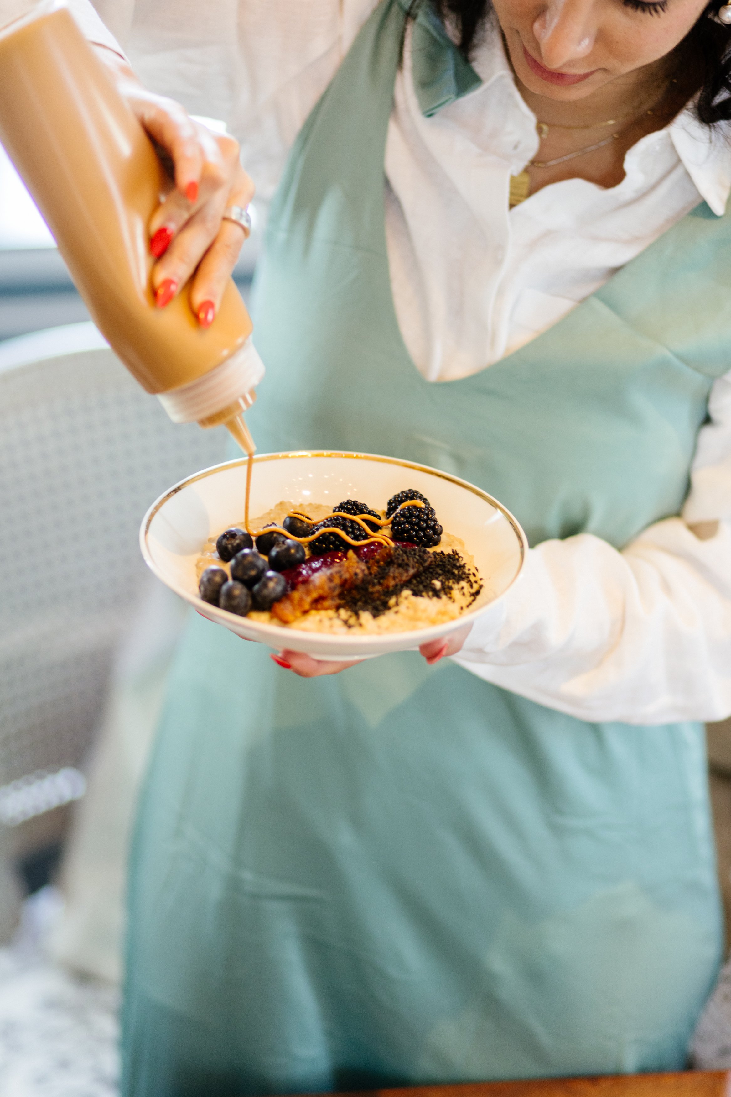 Une femme verse du caramel sur un bol de panna cotta garnie de fruits noirs et de crème