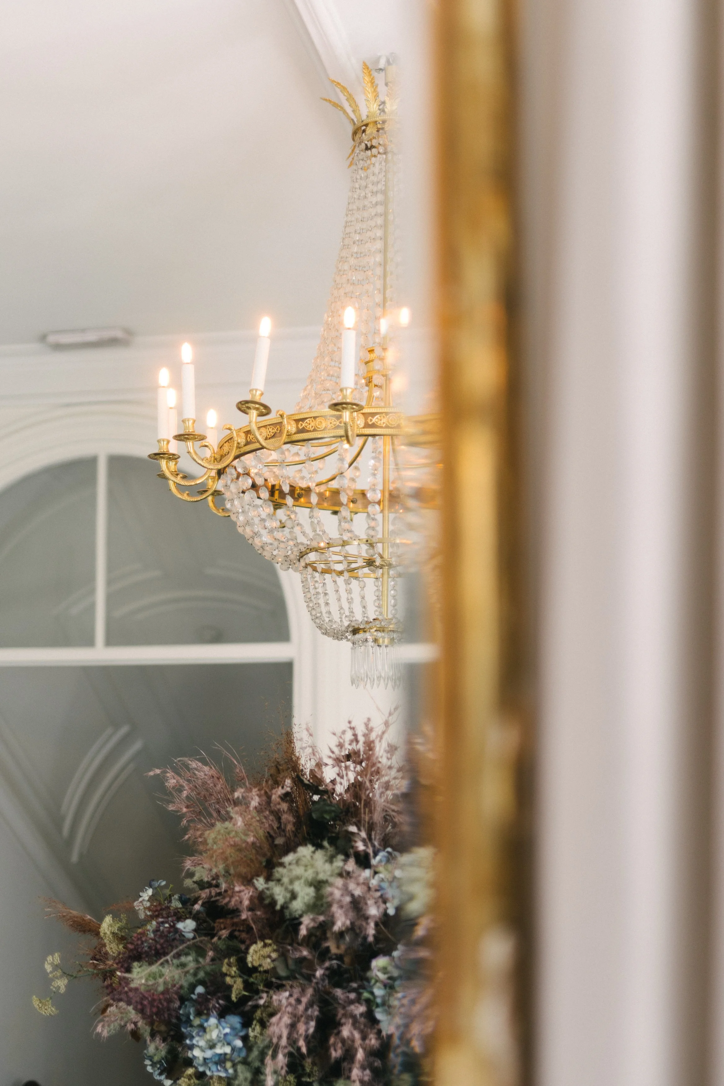Un lustre en cristaux et or suspendu au plafond dans une salle élégante, avec un grand bouquet de fleurs séchées en dessous.