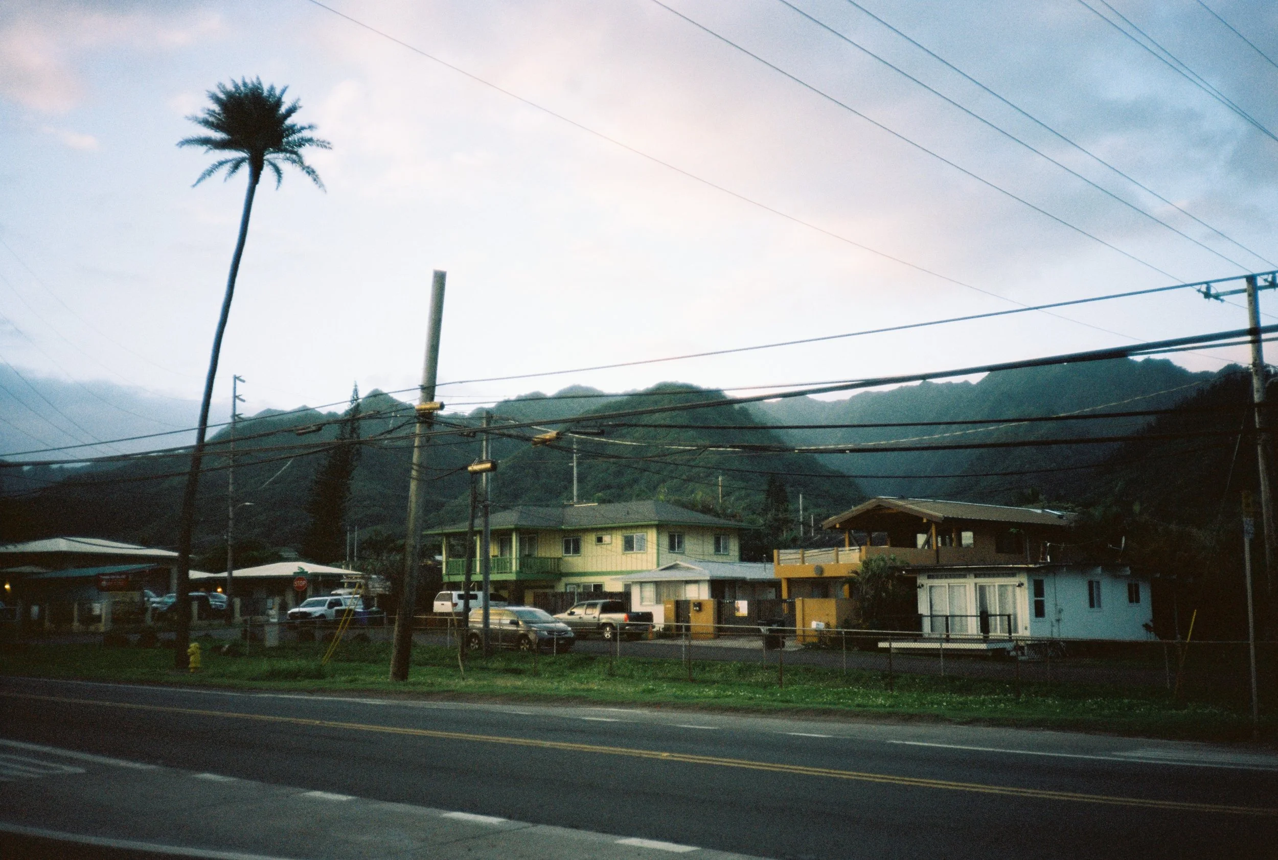 Maisons colorées avec voiture devant, montagne en arrière-plan, pylônes électriques, route au premier plan.