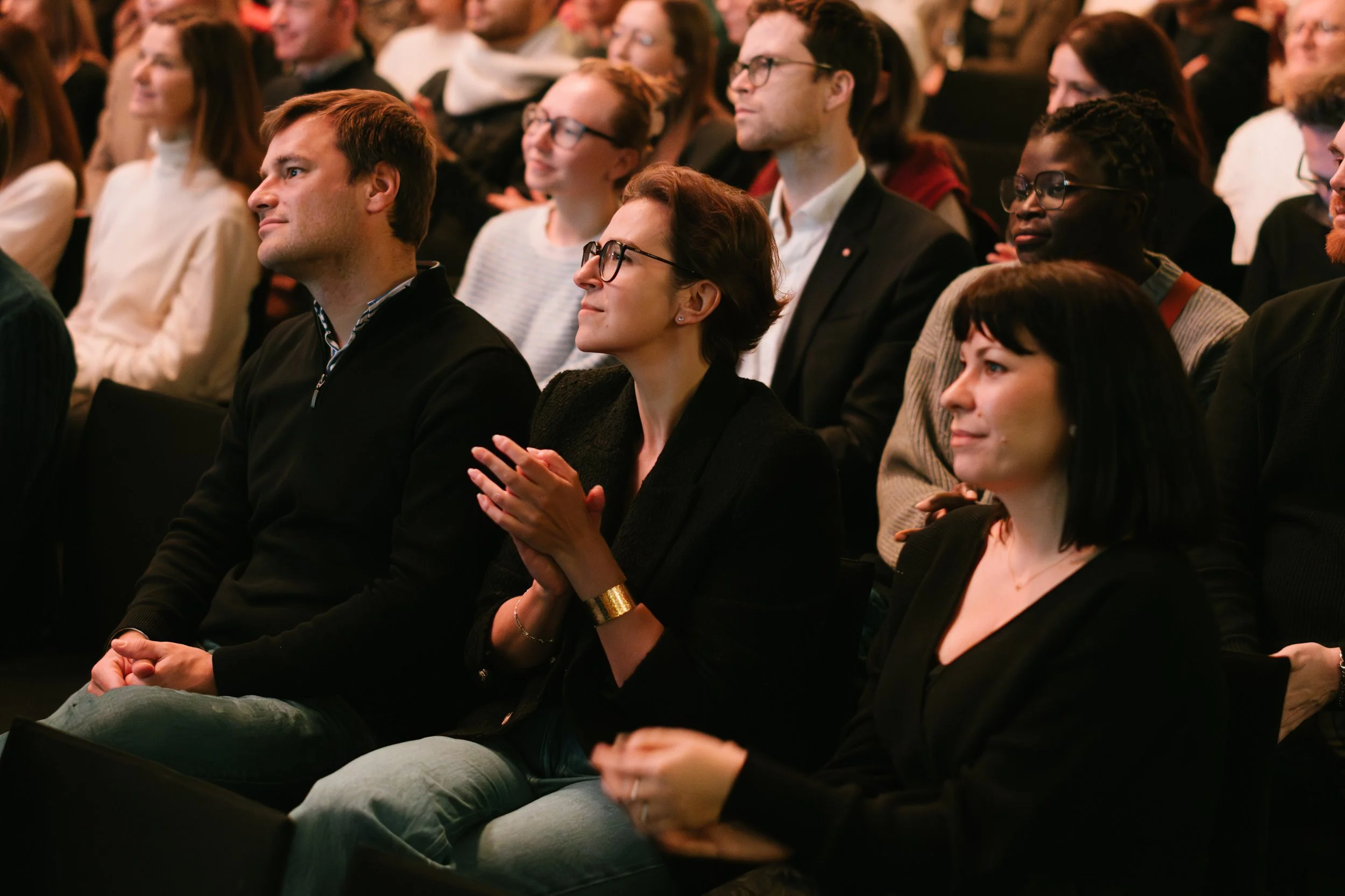 Groupe de personnes assises dans une salle de conférence, écoutant attentivement et applaudissant lors d'un événement.