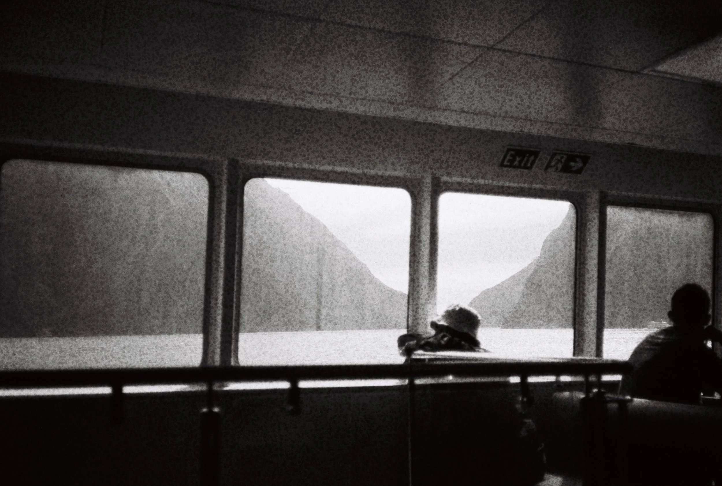 Une scène en noir et blanc à l'intérieur d'un ferry ou bateau, avec deux personnes assises près des fenêtres, regardant un paysage avec des montagnes et un corps d'eau à l'extérieur.