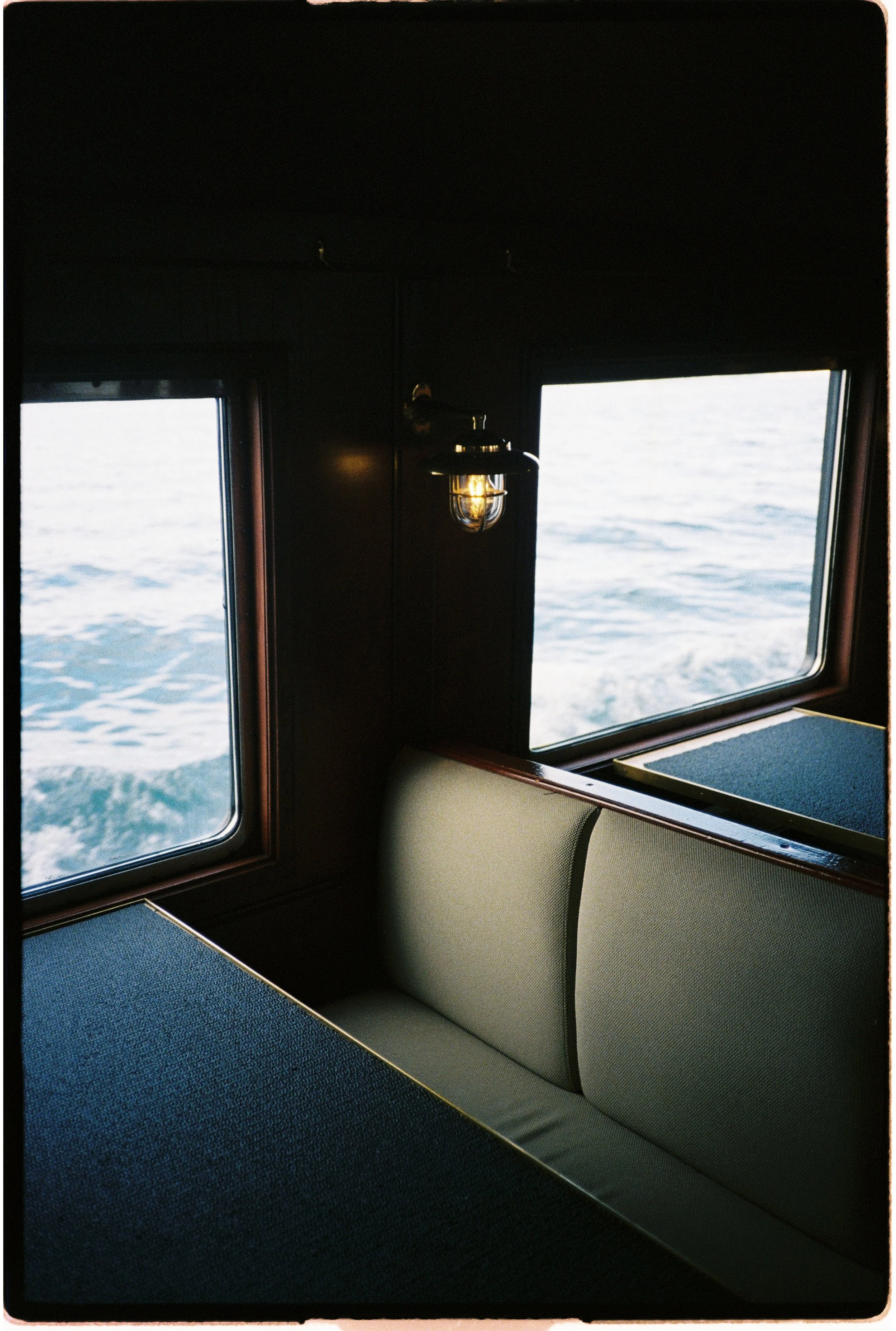 Intérieur d'une cabine de bateau avec deux fenêtres, vue sur la mer, et un lampadaire vintage. Meubles en tissu vert.