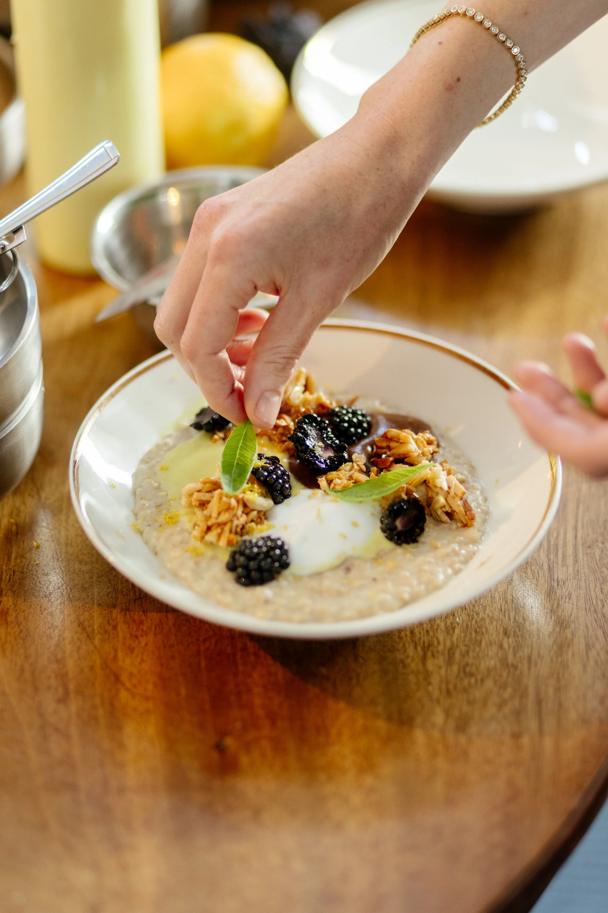 Une main décore un plat de porridge ou d'avoine avec des mûres noires, des noix, des feuilles vertes et un œuf poché, sur une table en bois.