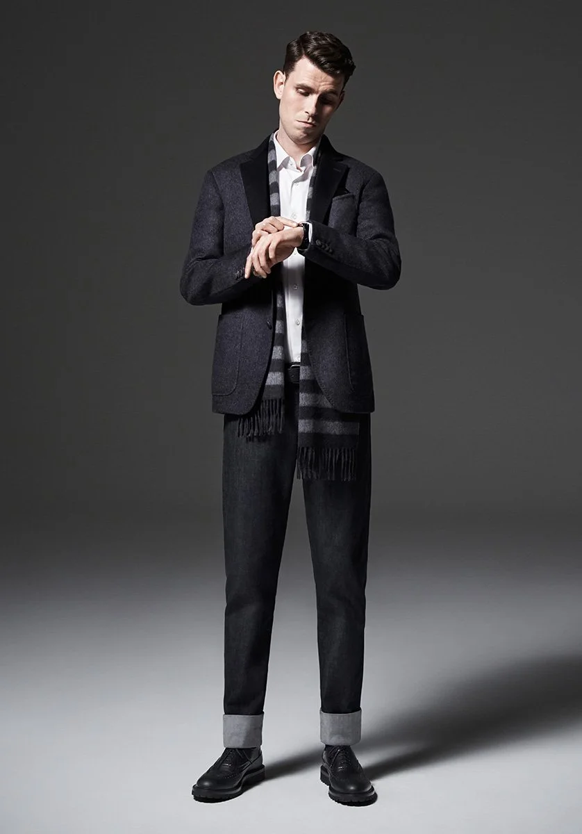 Young man in formal attire checking his watch, standing against a plain gray background.