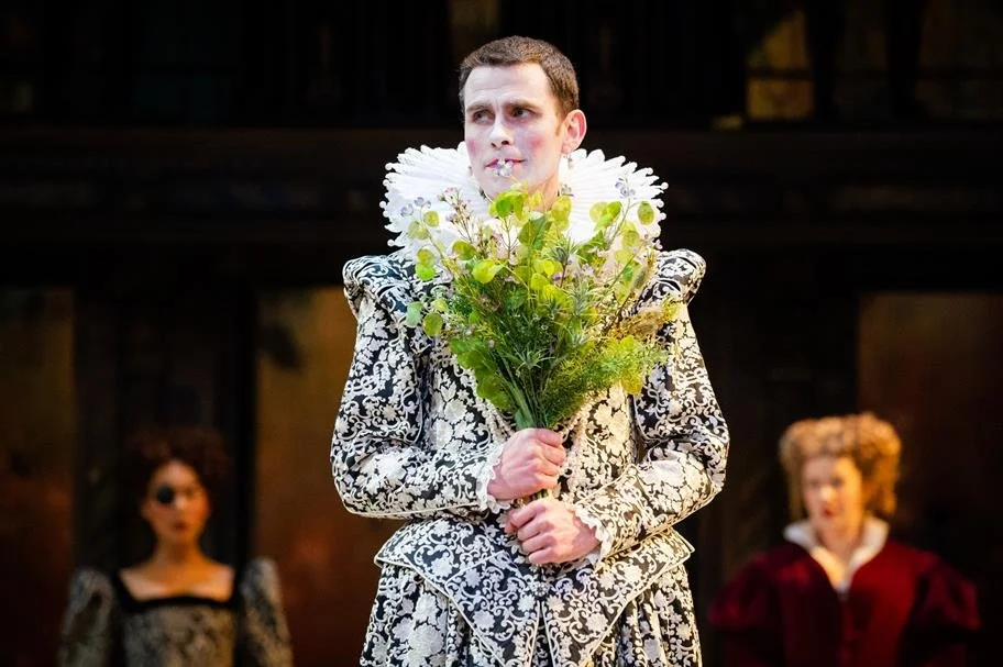 A young man dressed in an ornate, historical costume holding a bouquet of flowers, with two women in period dresses in the background.