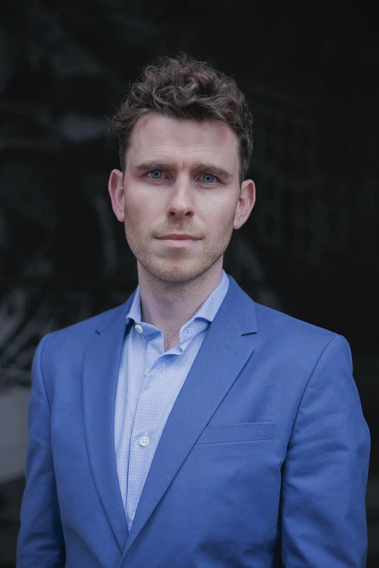A young man with curly brown hair and blue eyes wearing a blue suit jacket and a light blue dress shirt, standing outdoors against a dark background.