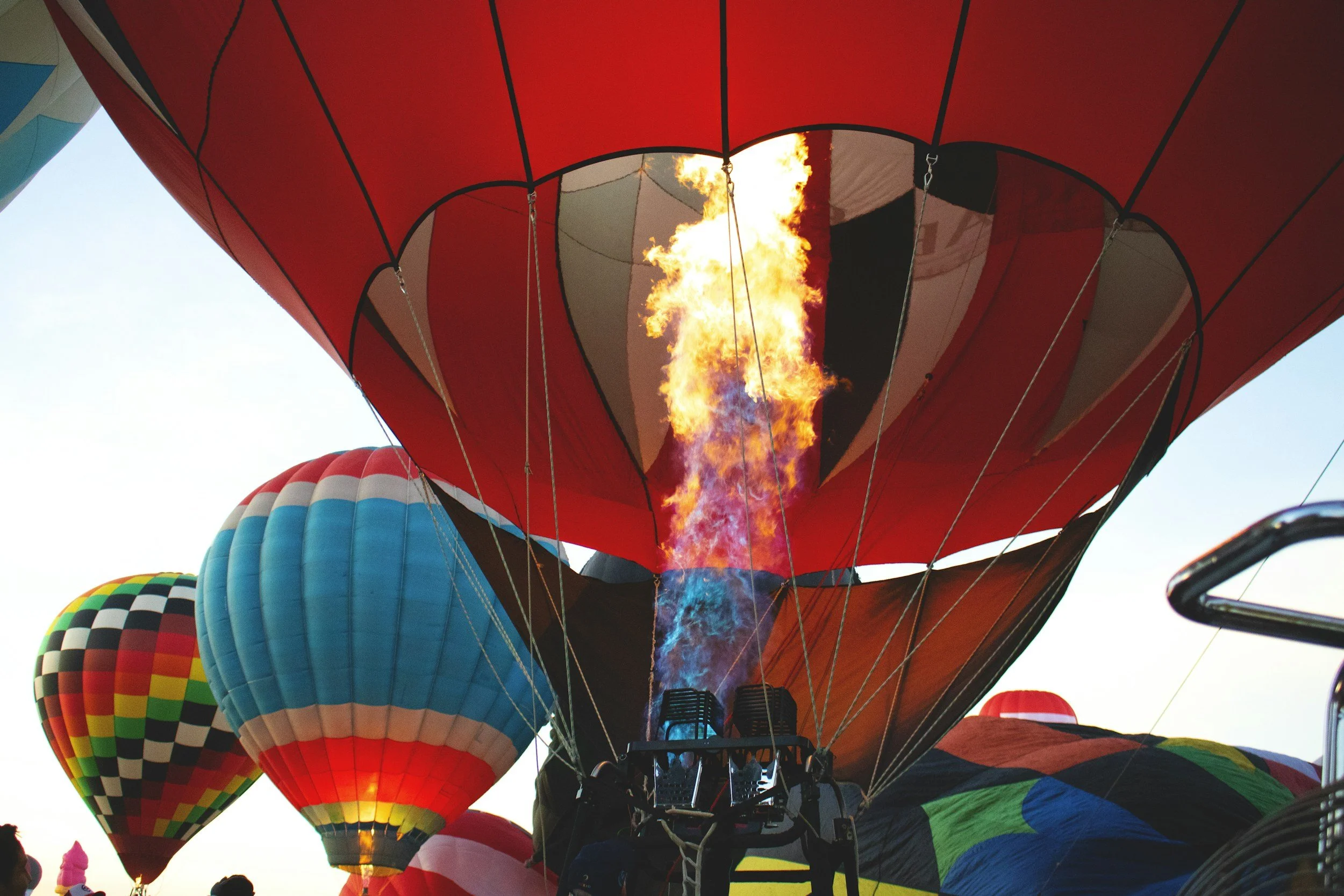 Colorful hot air balloons, with one inflating with flames coming from the burner, against a clear sky.
