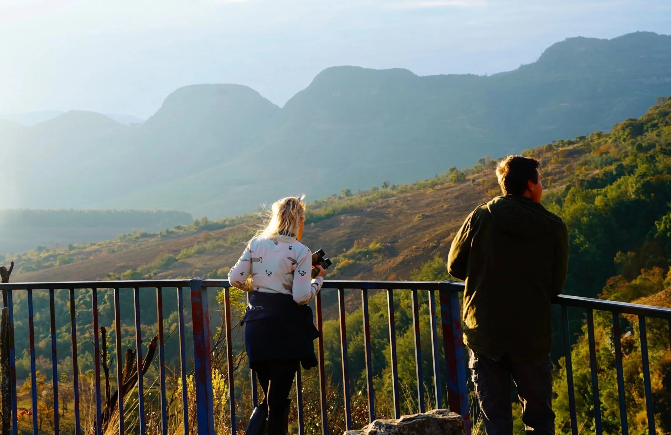 Two people stand on a viewing platform overlooking a scenic mountain landscape during sunset.