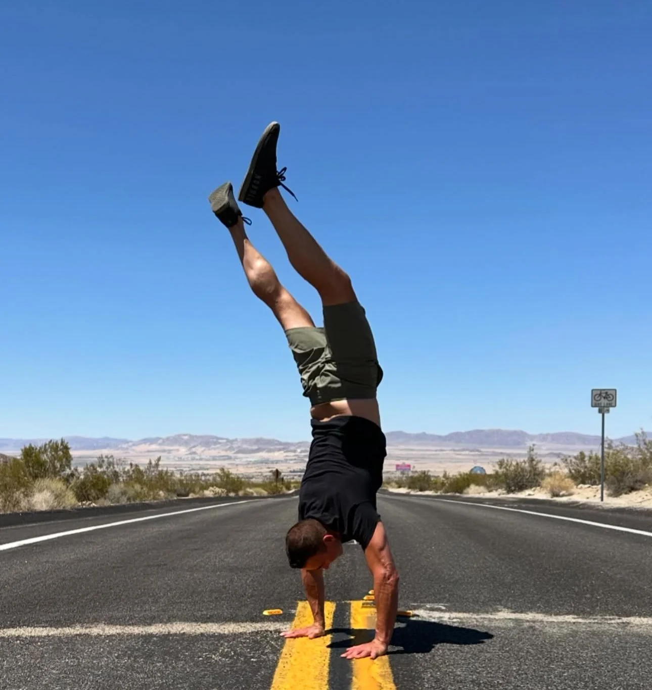 A man doing a handstand on a deserted road with a mountain landscape in the background and a bicycle lane sign visible.
