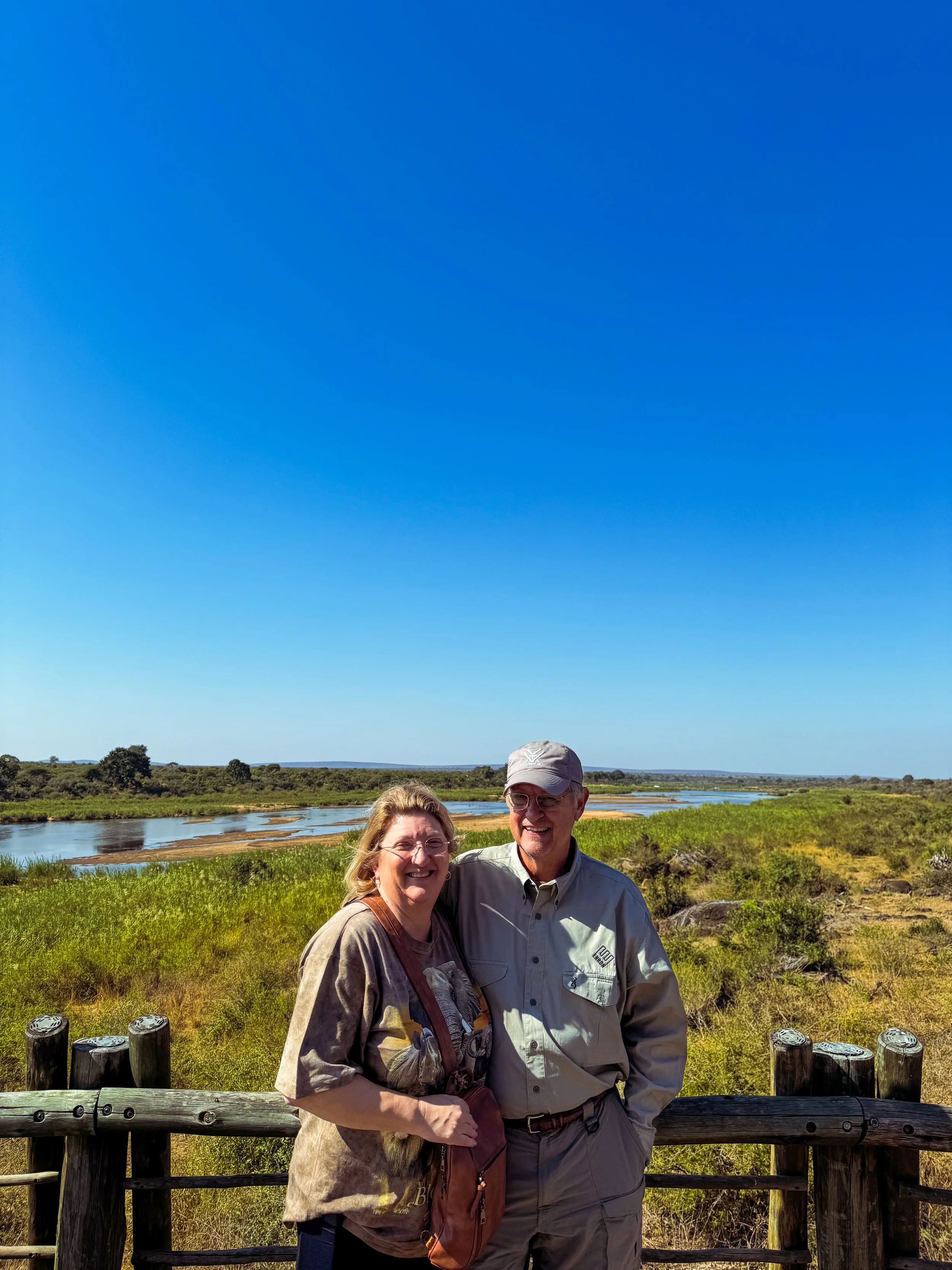 A smiling couple standing on a wooden railing in a natural landscape with a winding river and green vegetation under a bright blue sky.