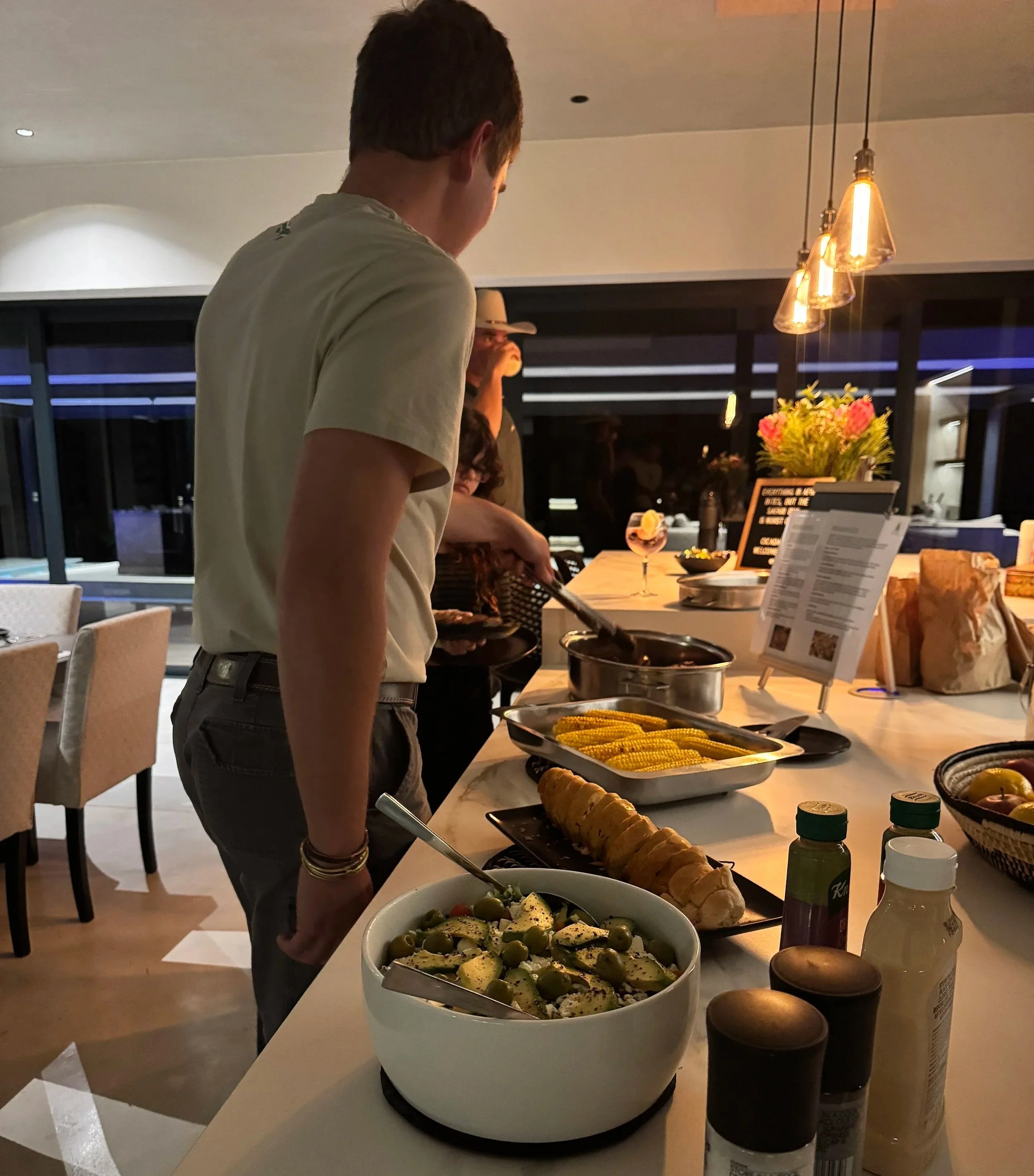 People serving food at a buffet in a modern, well-lit dining area with a view outside at night.