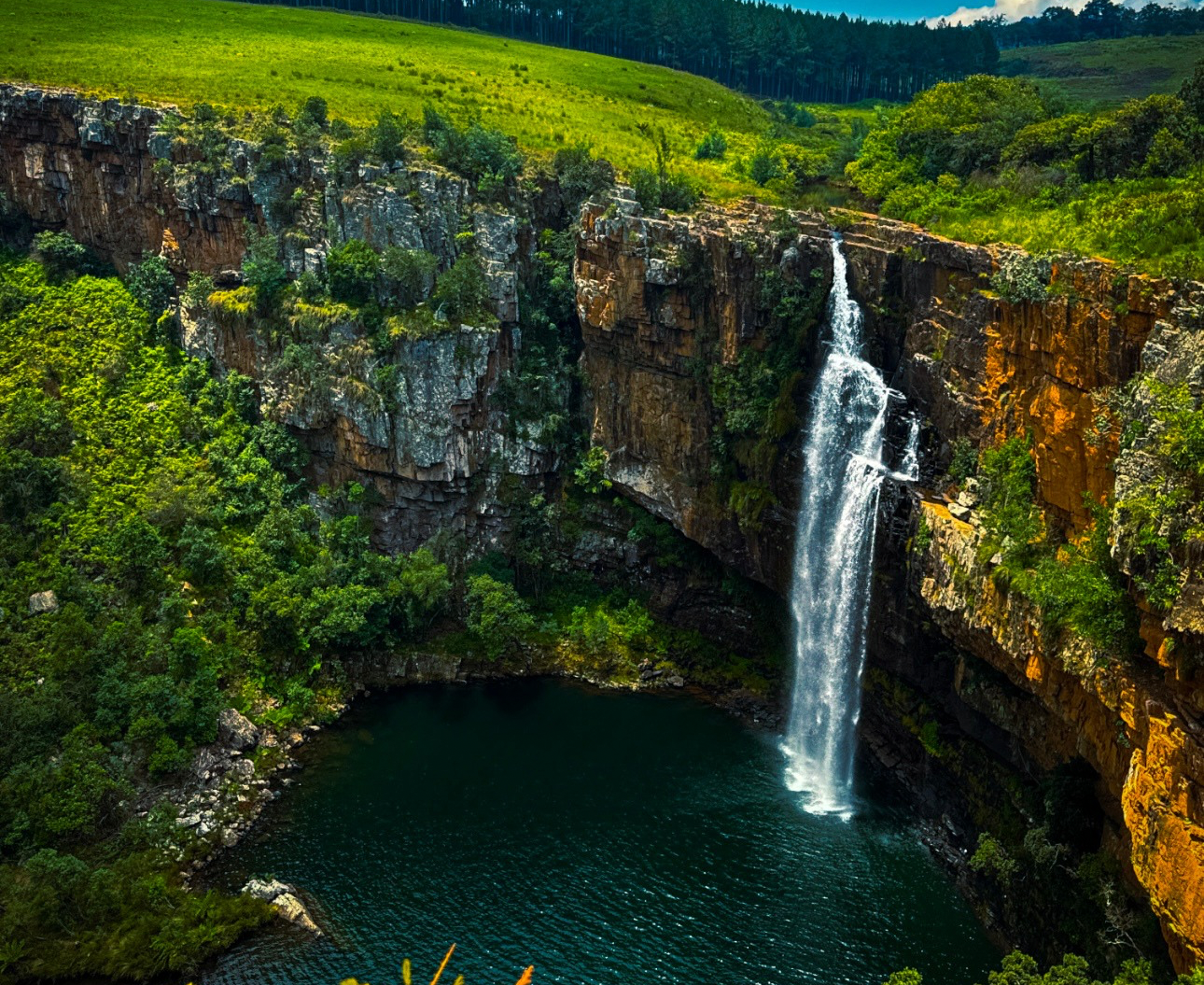 A waterfall flowing down a rocky cliff into a dark green pool surrounded by lush green trees and grass.
