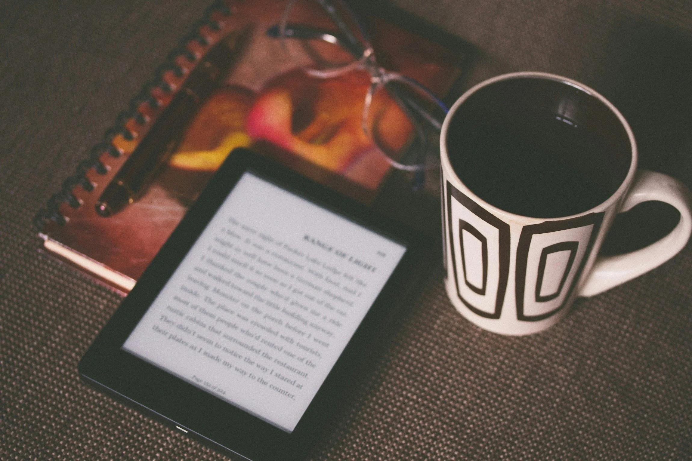 A tablet displaying an eBook, a spiral-bound notebook with a pen, a cup of black coffee, and a pair of eyeglasses on a textured surface.