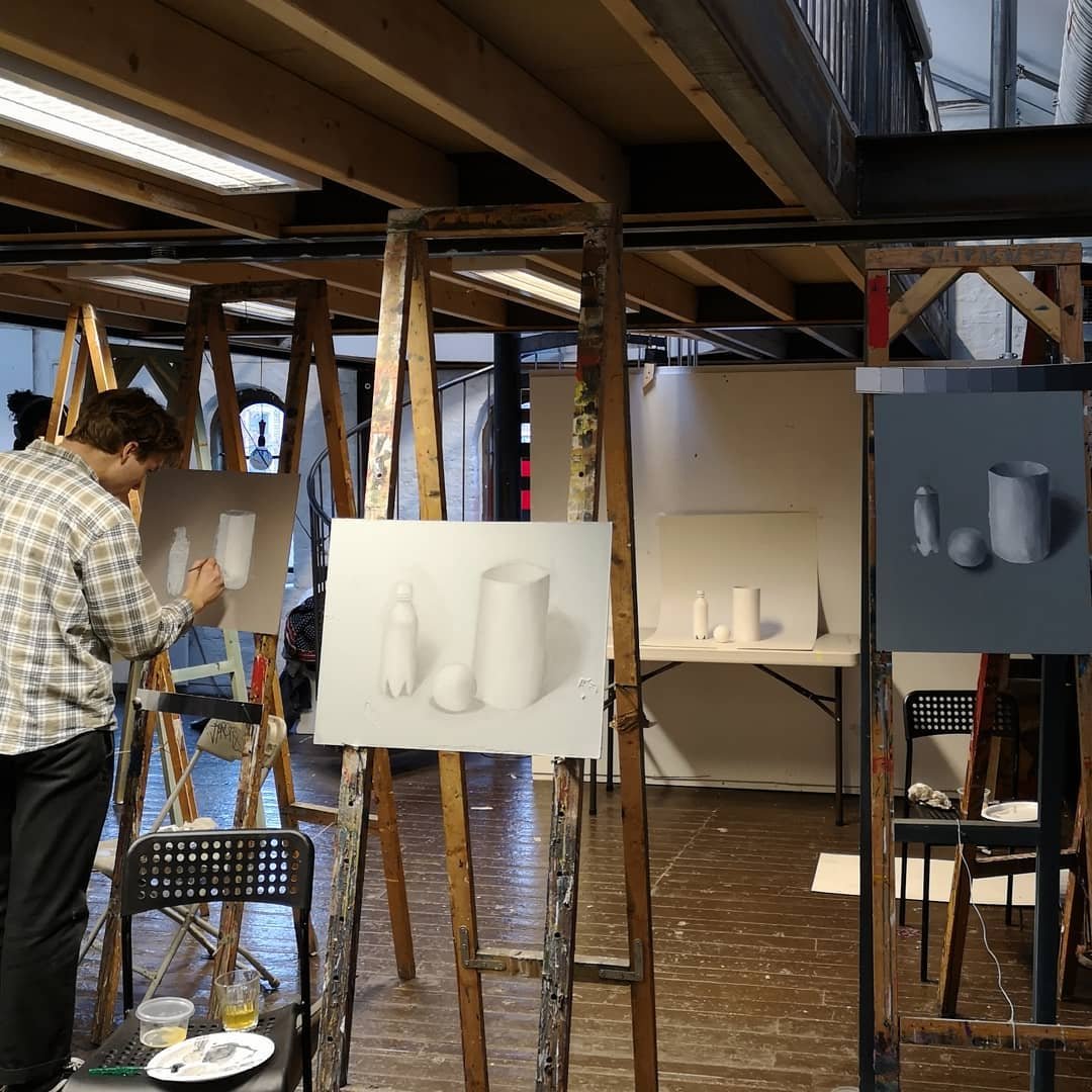 En kunstner maler på et staffeli i et rom med flere andre staffelier, der flere bilder av hvite objekter som flasker og baller er utstilt.